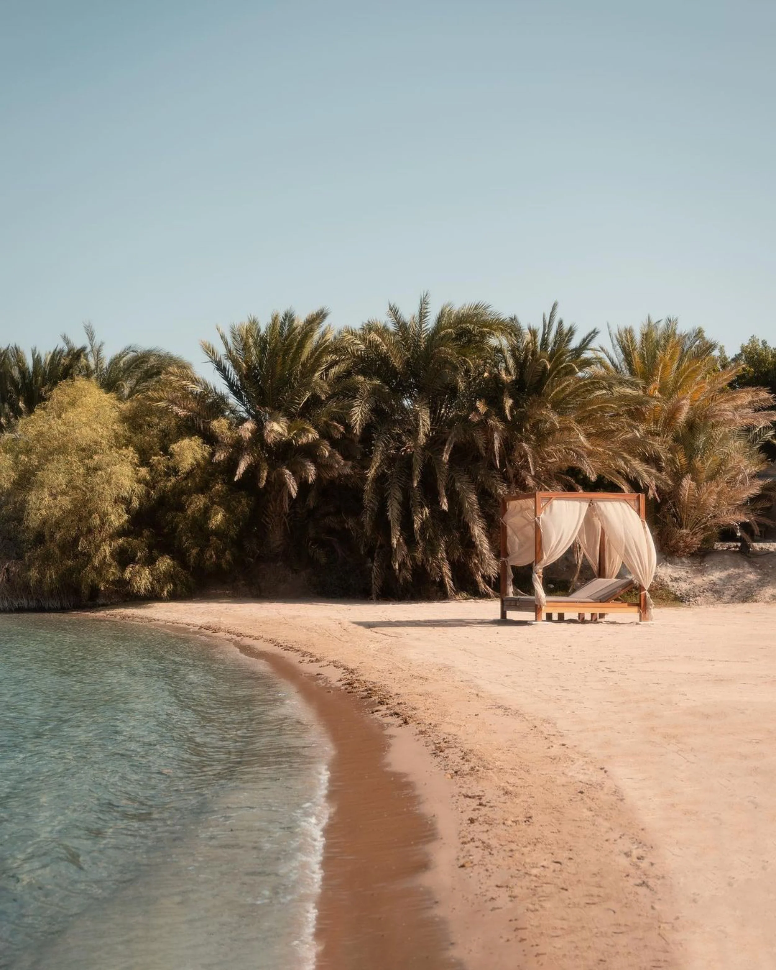 Beach with clear water, sandy shore, lush green trees, and a cabana with curtains