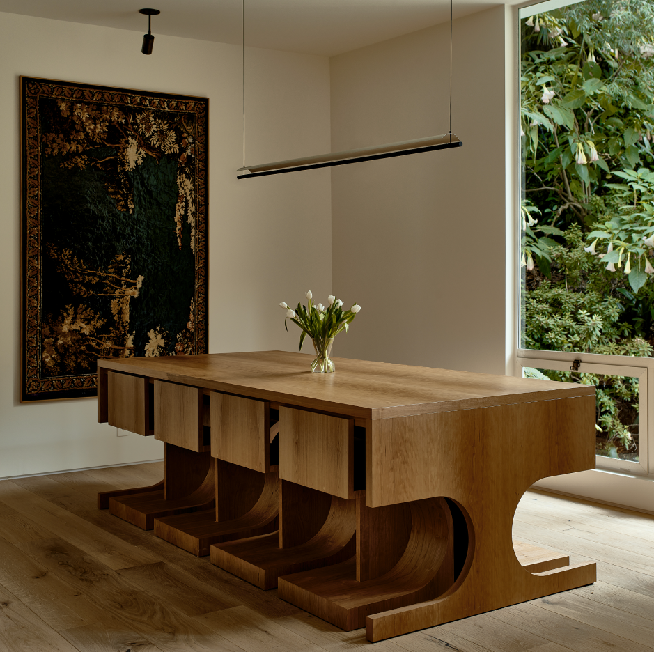 A minimalist dining room with a wooden table and four matching chairs, a vase of white tulips on the table, a large window with greenery outside, a dark tapestry on the wall, and a modern linear pendant light