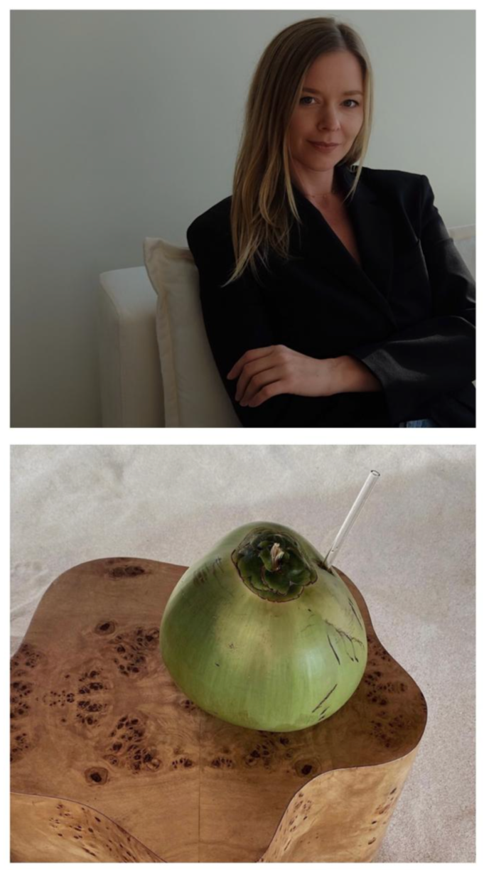 A woman with long blonde hair wearing a black shirt, sitting on a white couch with her arms crossed, smiling gently. A green coconut with a straw is placed on a wooden side table in front of her.