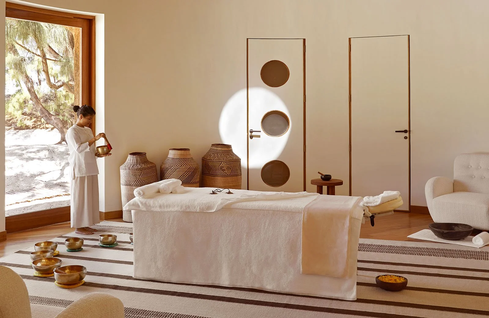 A woman in white clothing standing in a peaceful, minimalist massage or spa room with a massage table, decorative woven baskets, and natural light from a large window showing an outdoor scene with a tree.