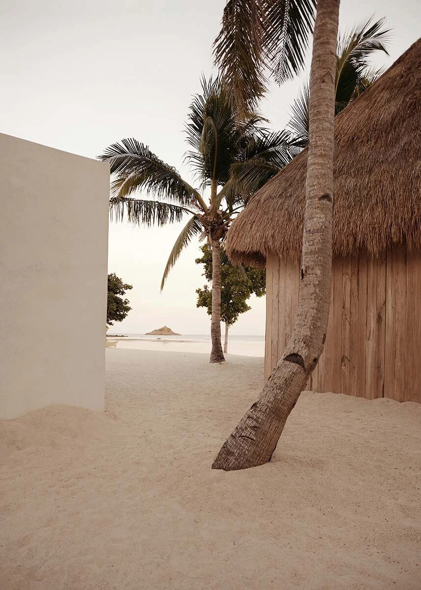A sandy beach with palm trees, a thatched hut, and an island in the distance.