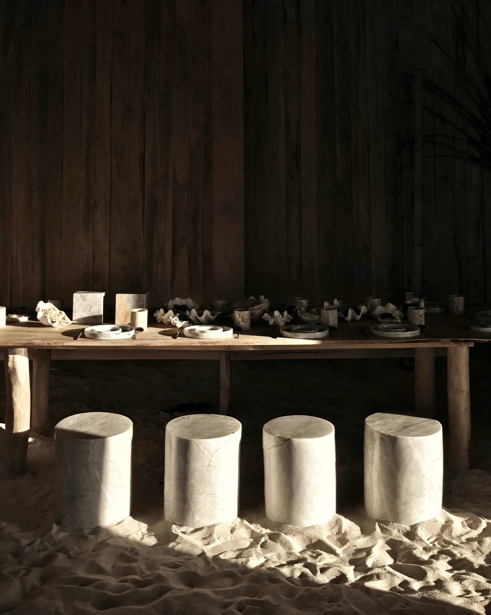 Outdoor space at night with a wooden fence, round white seats on a sandy ground, and objects on a wooden table.