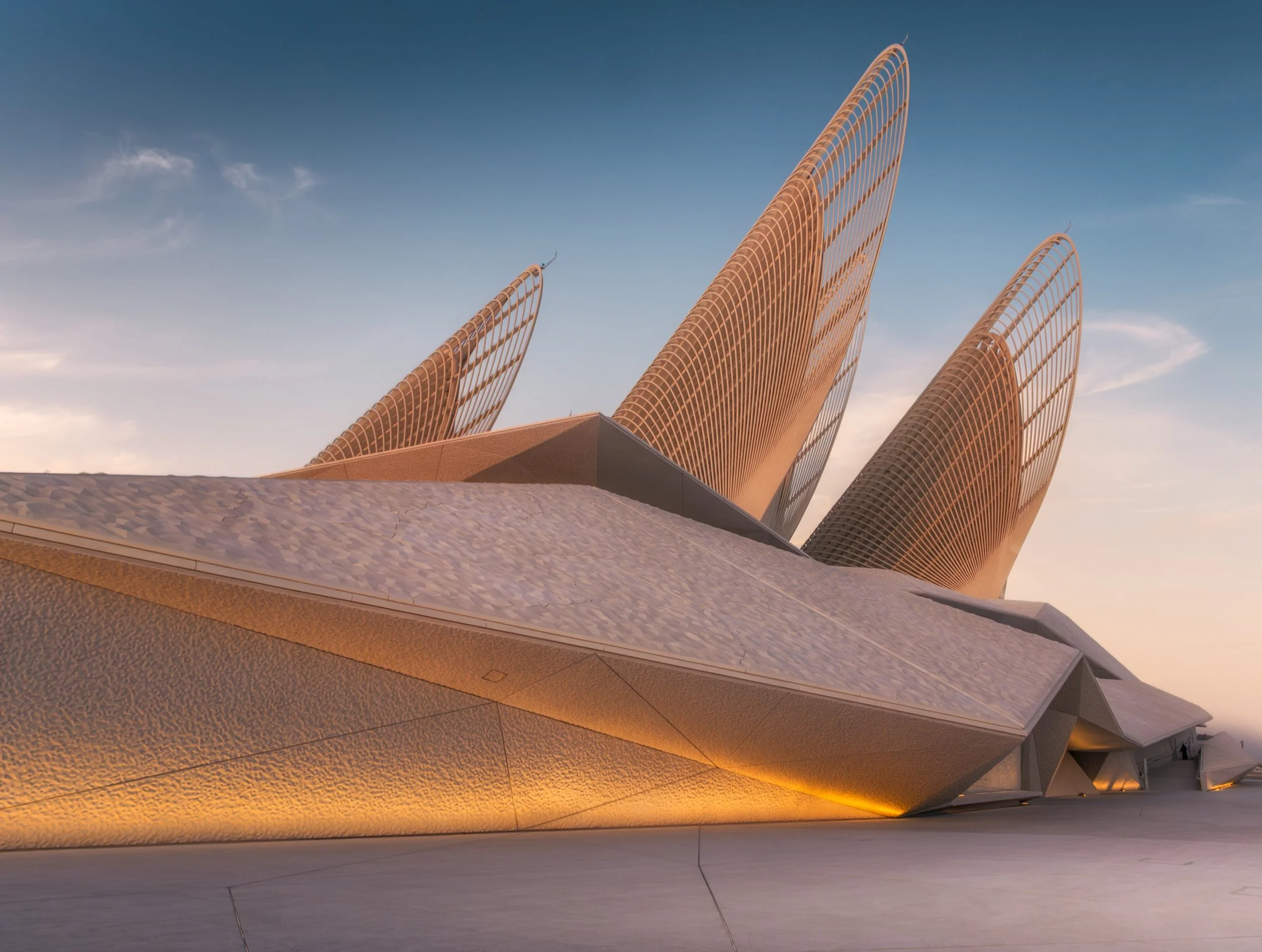 Modern architectural building with curved, sail-like structures under a clear blue sky during sunset.