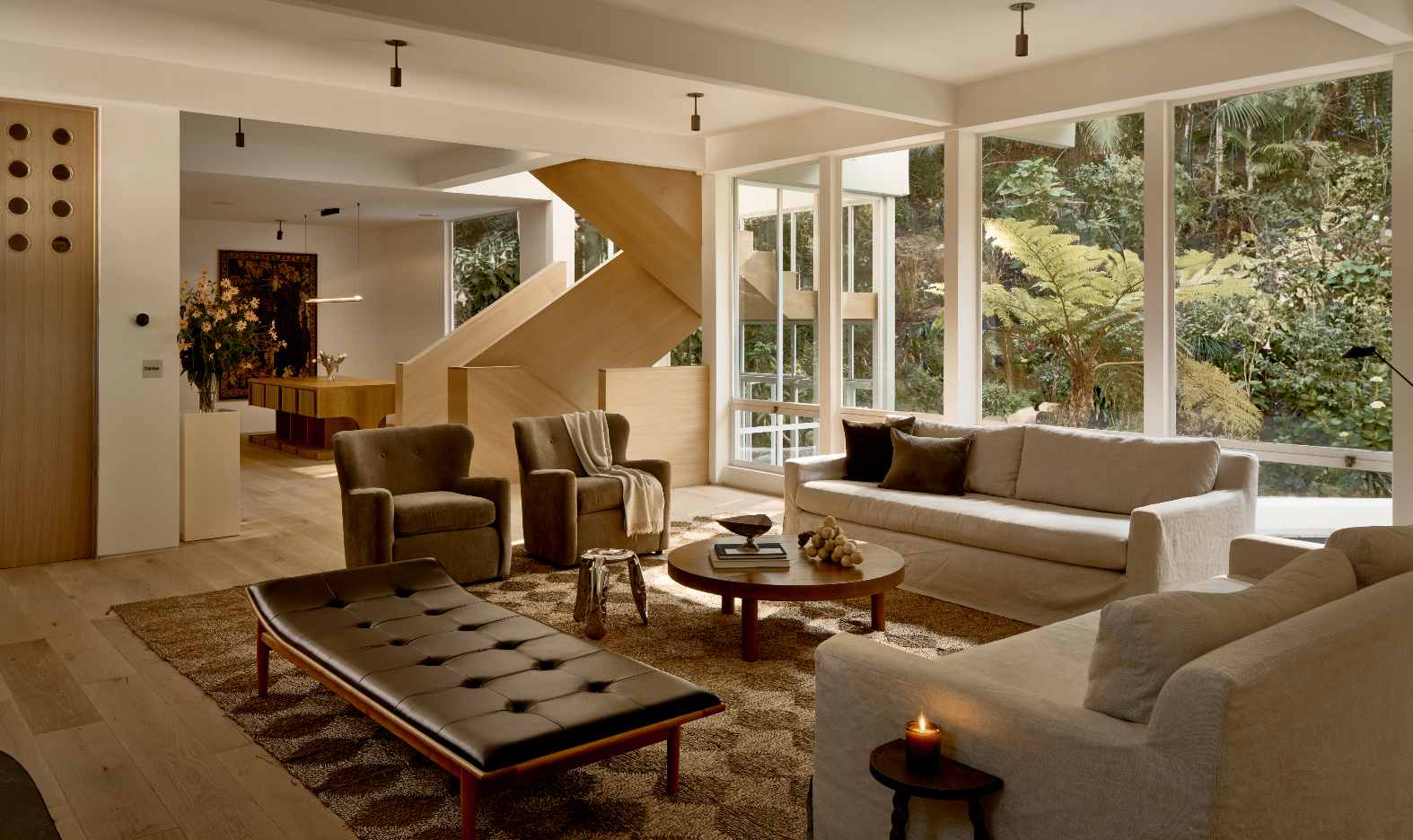 Living room with beige sofas, armchairs, wooden coffee table, and large windows overlooking greenery.
