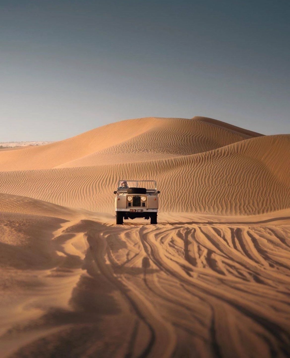 A white off-road vehicle driving through the desert sand dunes with tire tracks on the sand, under a clear blue sky.
