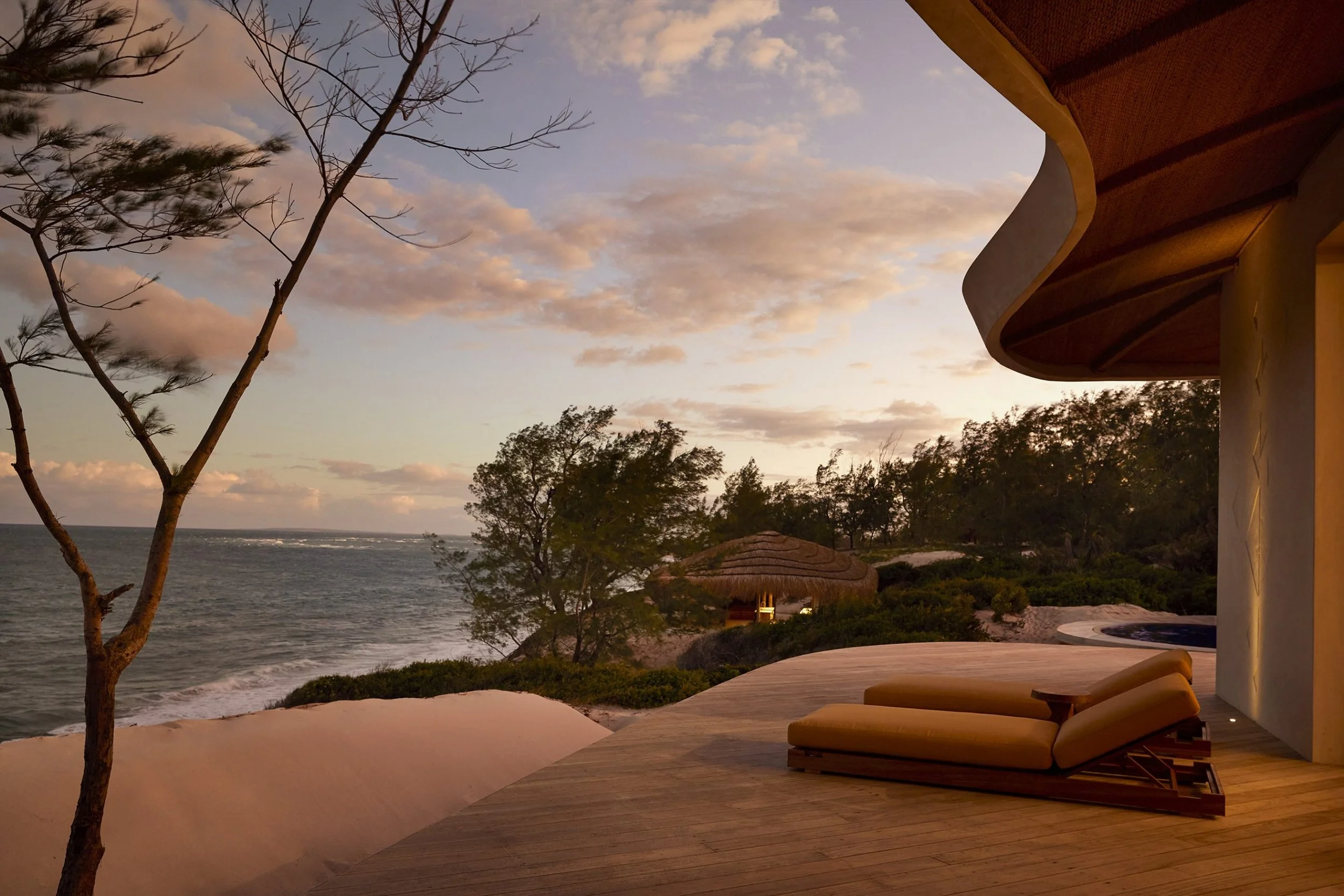 Beachside resort patio with cushioned lounge chairs, trees, ocean view, cloudy sky, and modern building with curved roof at sunset.