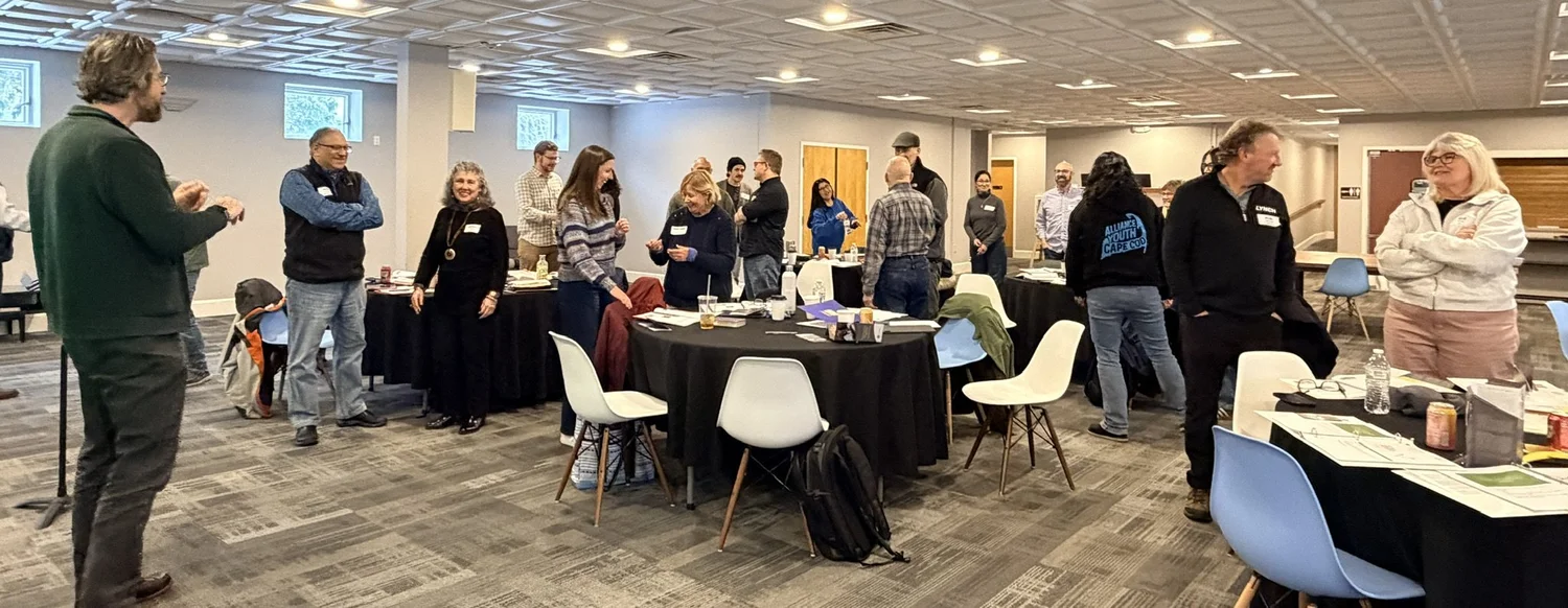 Pastors and leaders standing and talking around black-draped tables at an Alliance New England regional meeting, smiling as one leader addresses the room