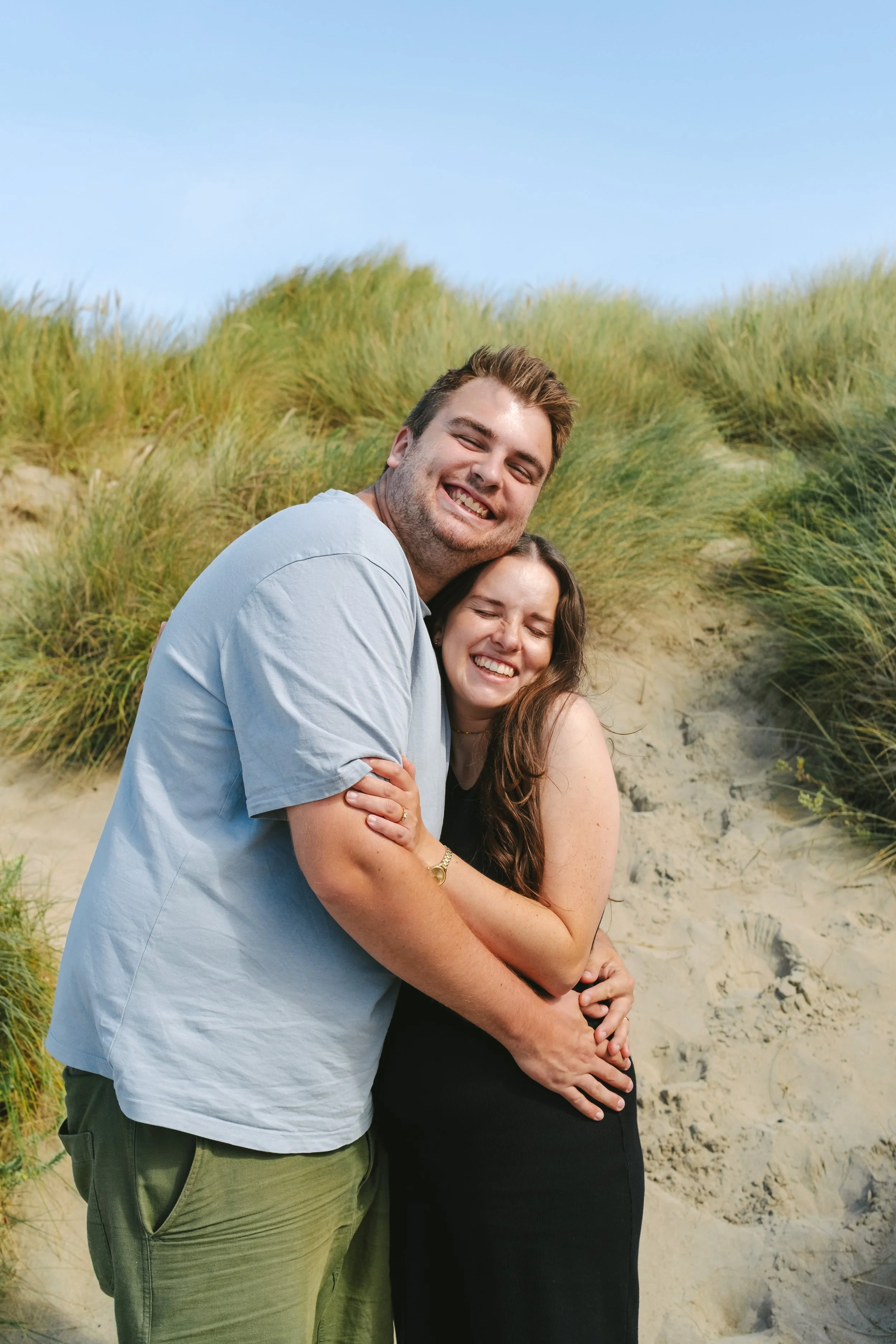 A happy couple, a man with light brown hair and a woman with brown hair, embracing and smiling at the beach, with dunes and green grasses in the background.