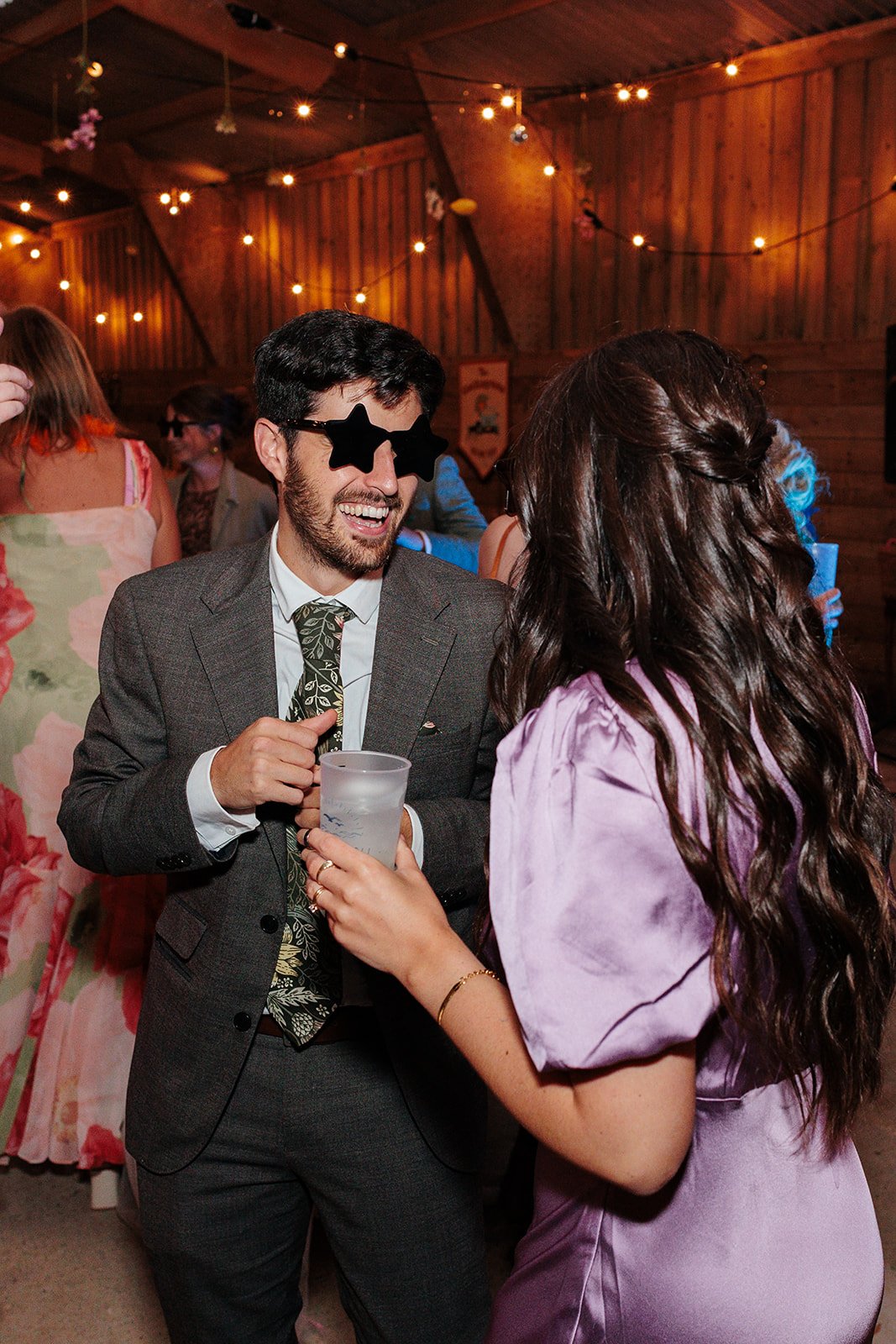 A man wearing star-shaped sunglasses and a suit, laughing and holding a drink, talking to a woman with long wavy hair in a lavender dress, at a party or celebration with string lights and wooden walls.