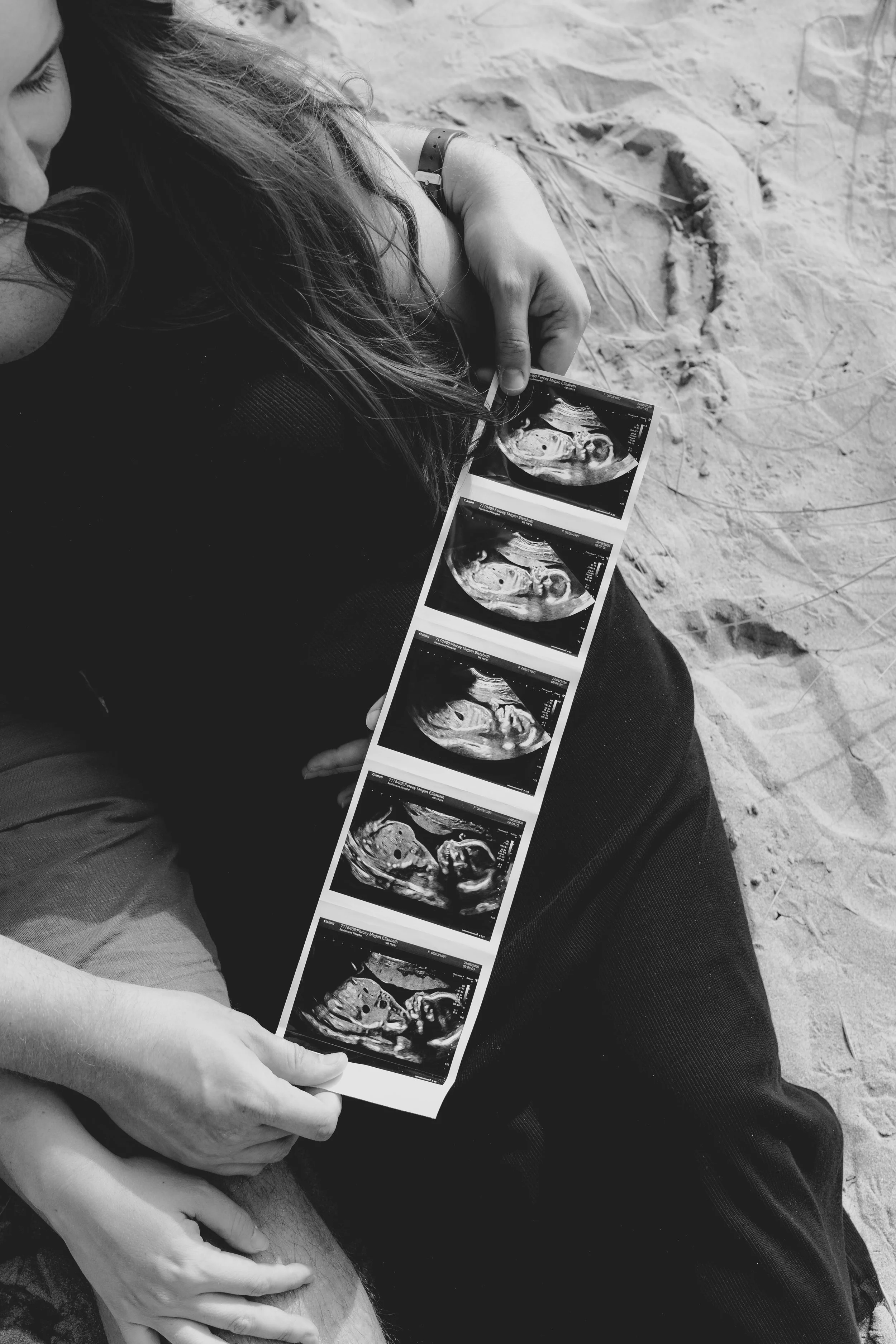 Black and white photo of a woman holding ultrasound images of a fetus, sitting on the sand, with another person's hand lightly touching her knee.