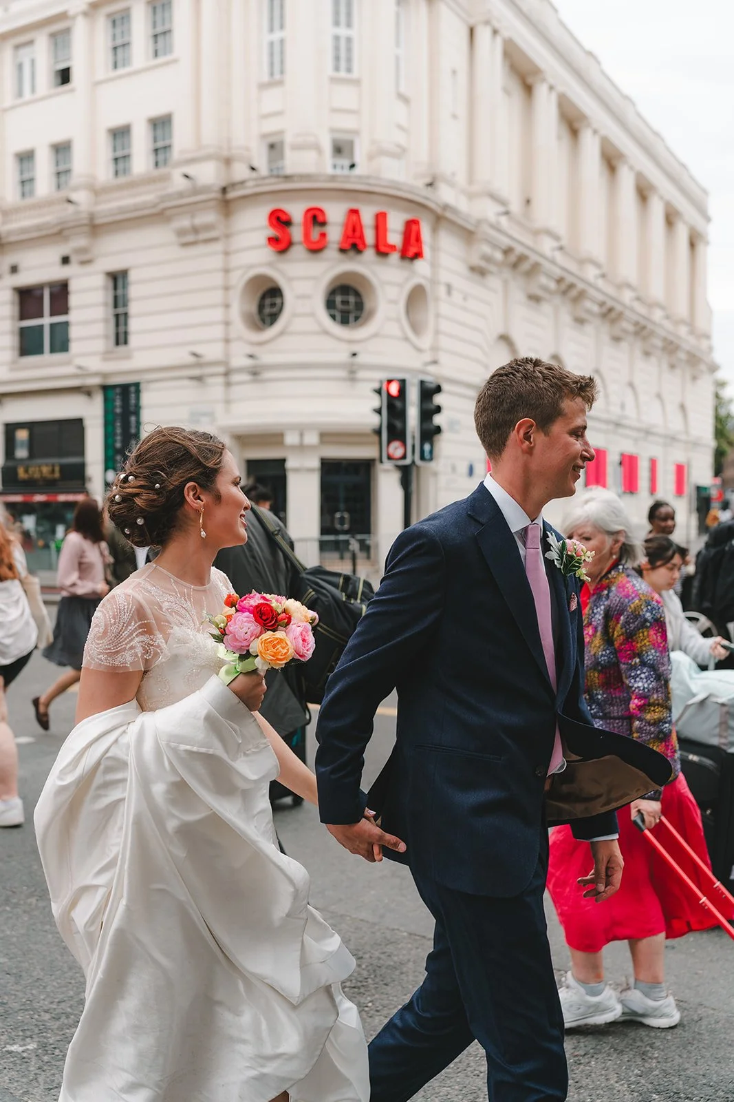 A bride and groom walking hand in hand on a city street during a wedding celebration, with the word 'SCALA' on a building in the background.
