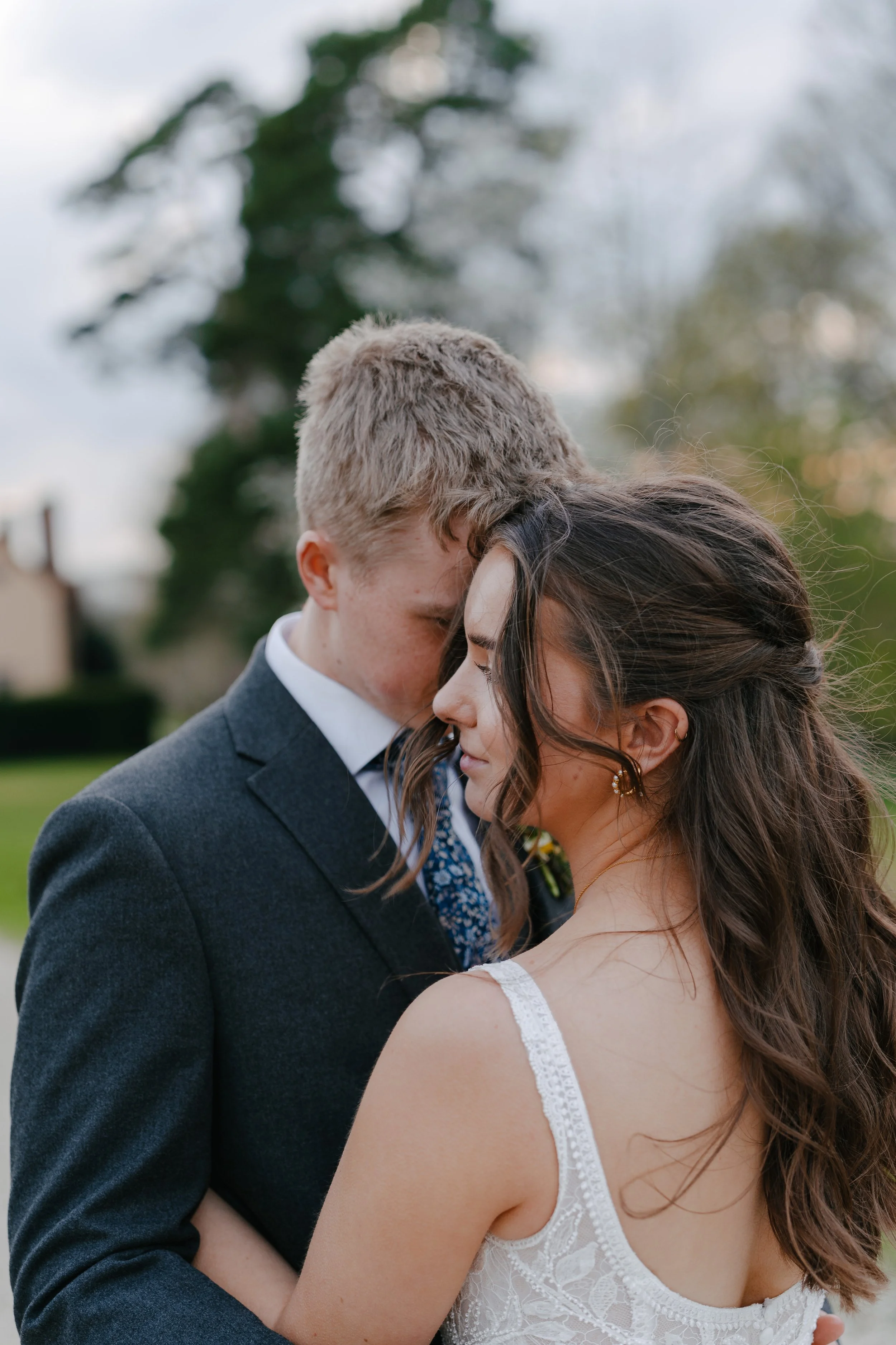 A couple, dressed in wedding attire, sharing an intimate moment outdoors with their foreheads touching and eyes closed, surrounded by natural scenery.