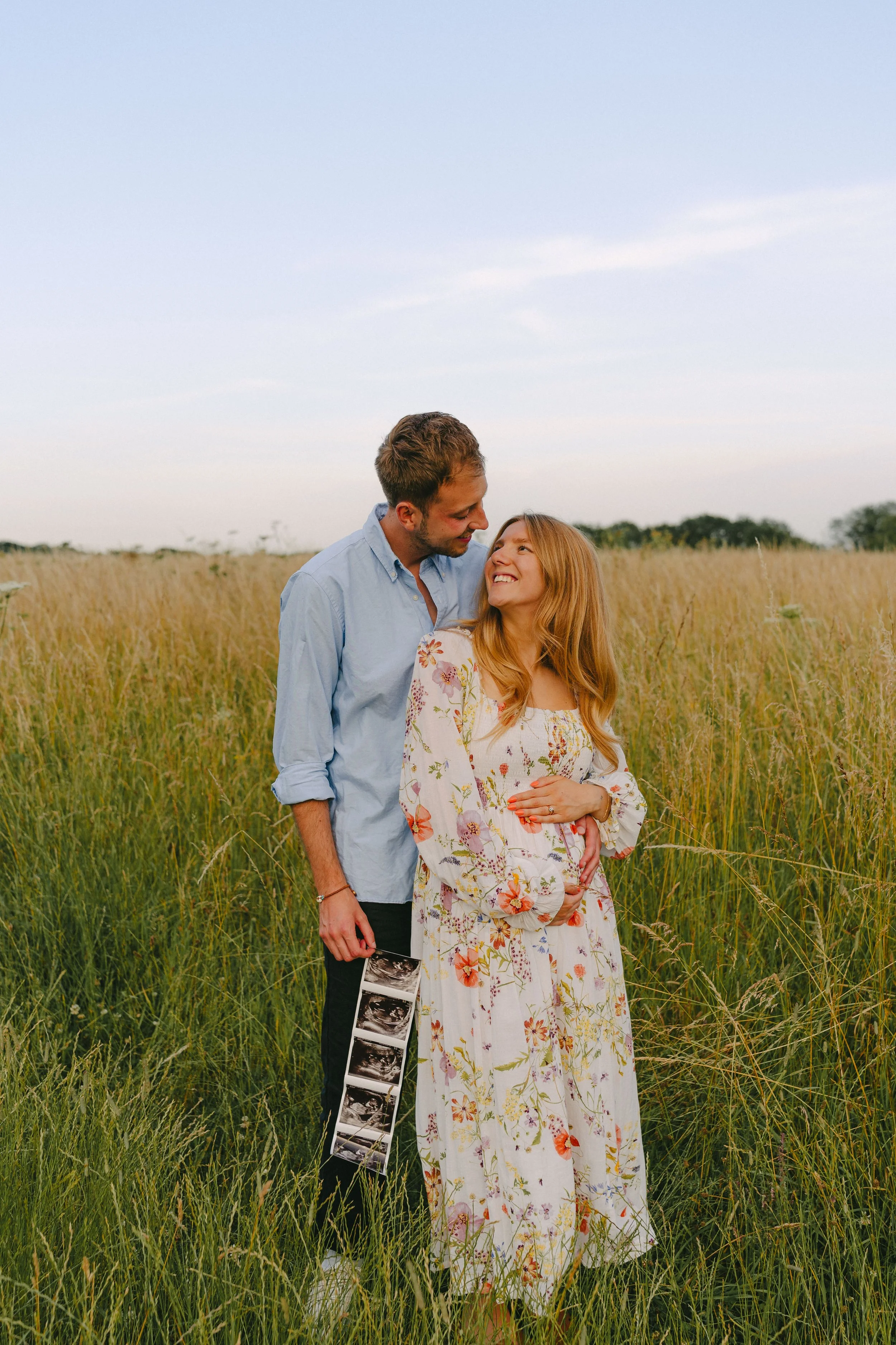 A couple in a field of tall grass, smiling and looking at each other. The man is holding ultrasound images, and the woman is wearing a floral dress.