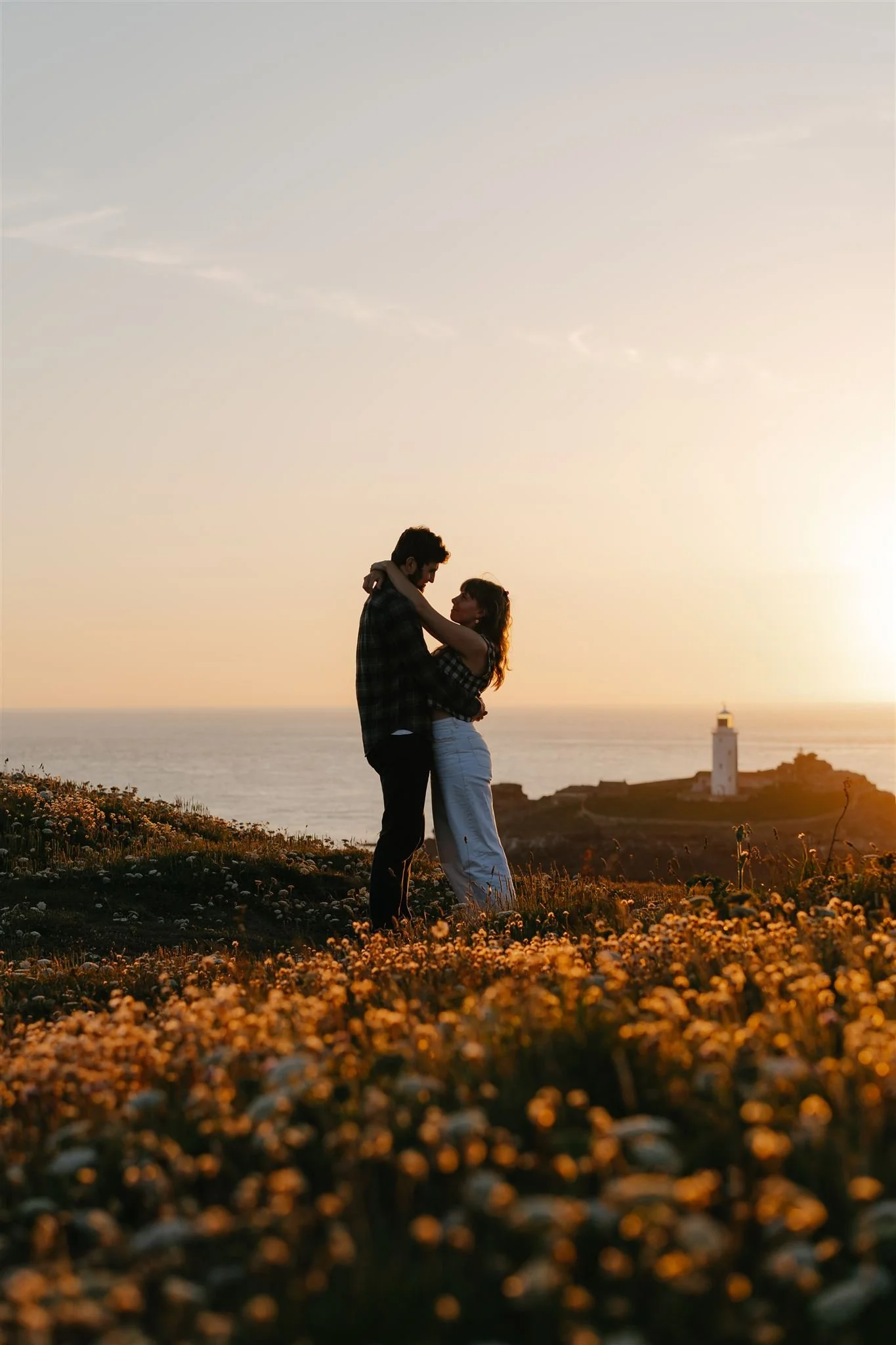 A couple embracing and kissing during sunset on a field of flowers near a lighthouse by the ocean.