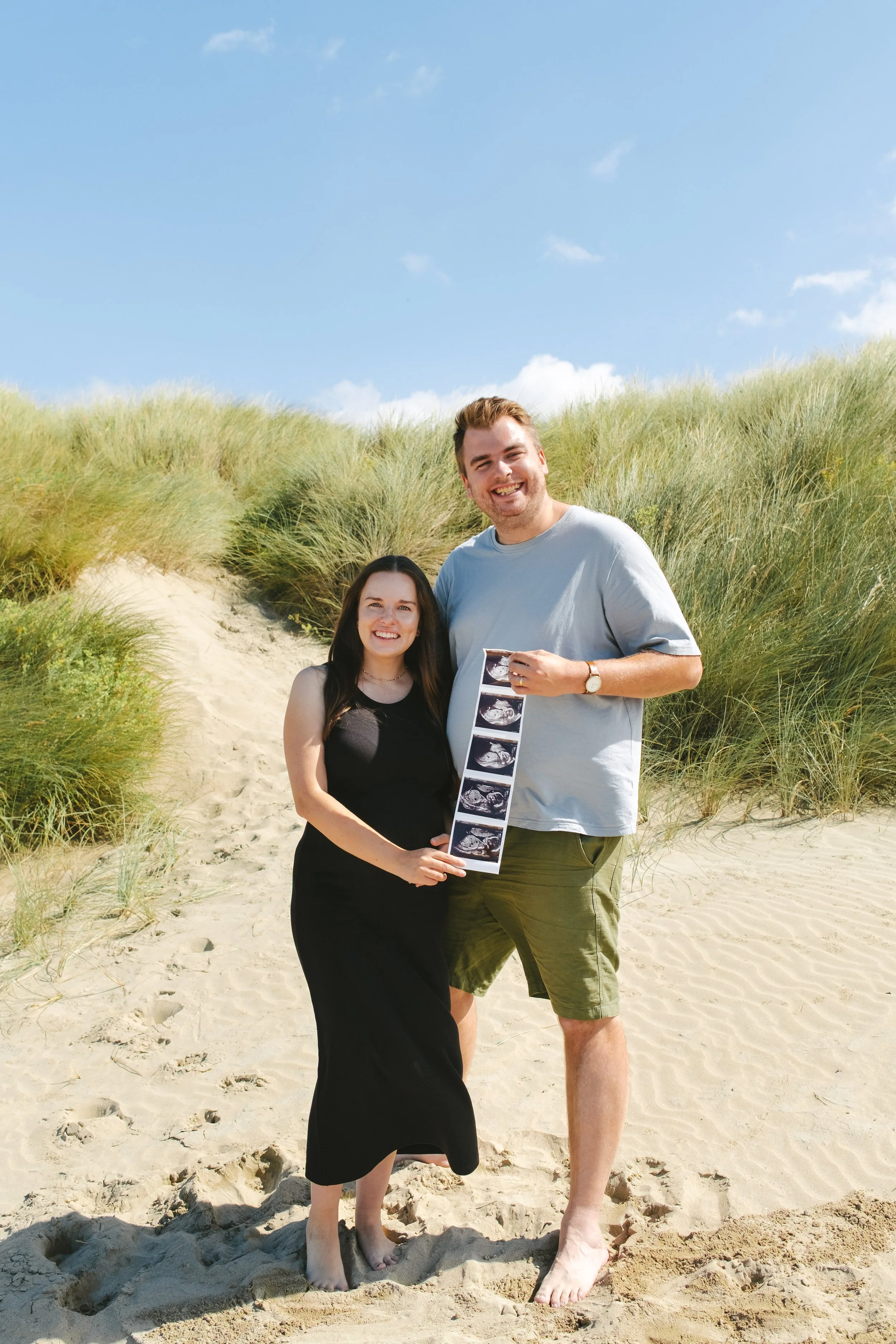 A smiling couple standing on a sandy beach with green dunes and a blue sky in the background. They are holding ultrasound photos.