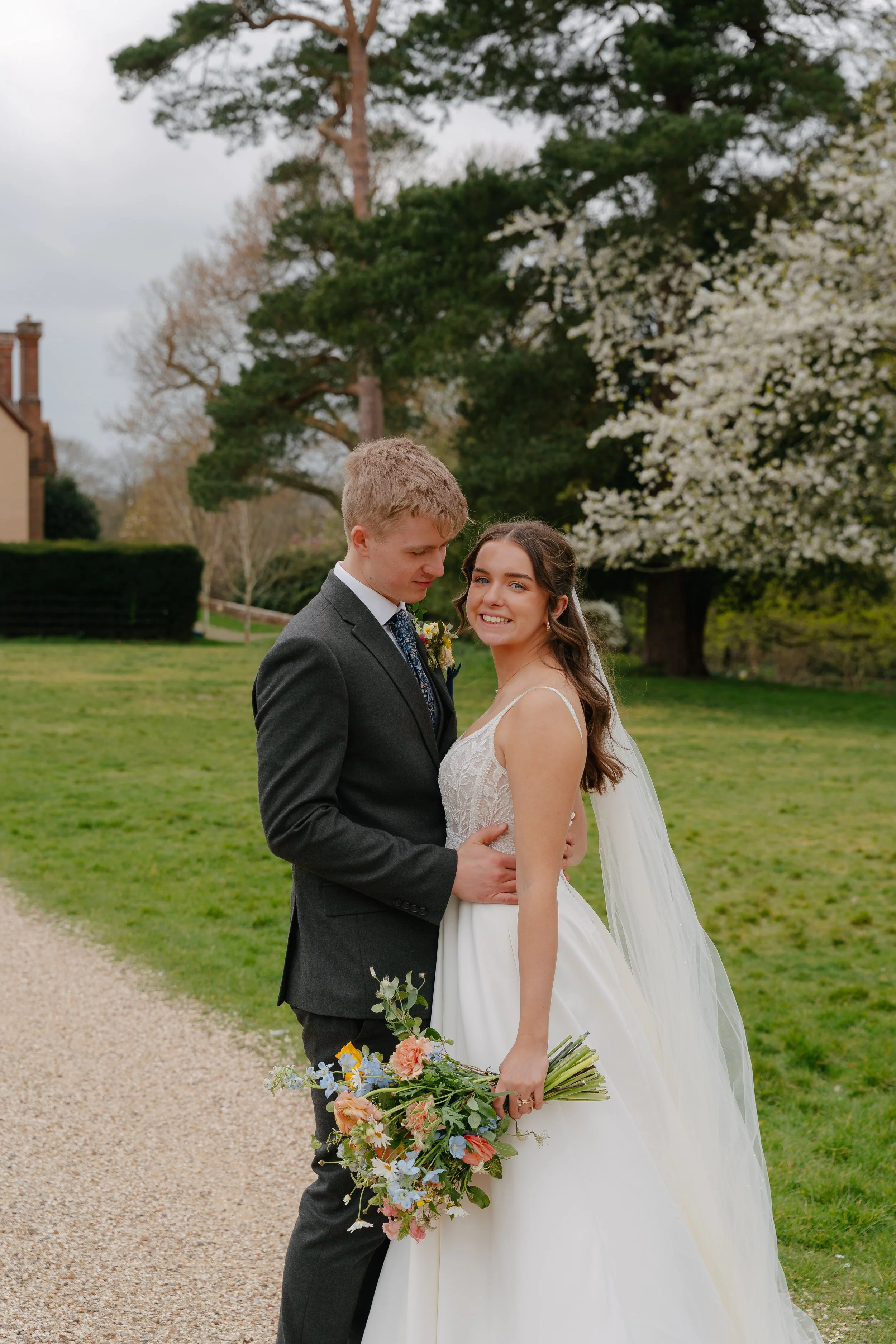 A bride and groom standing close outdoors on a green lawn, dressed in wedding attire, with blooming trees and a house in the background.