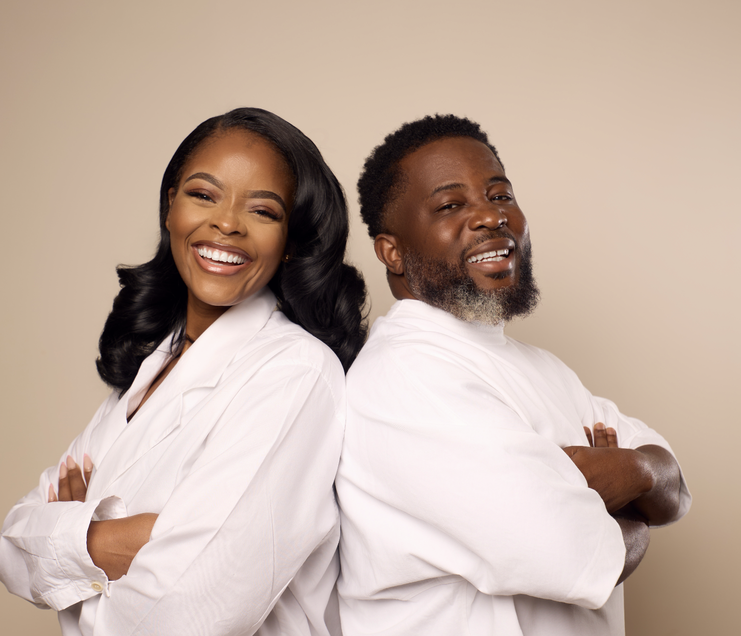 Gina Danso A smiling Black woman with long black wavy hair and a beard man with short curly hair, both wearing white shirts, standing back to back with arms crossed against a plain beige background.