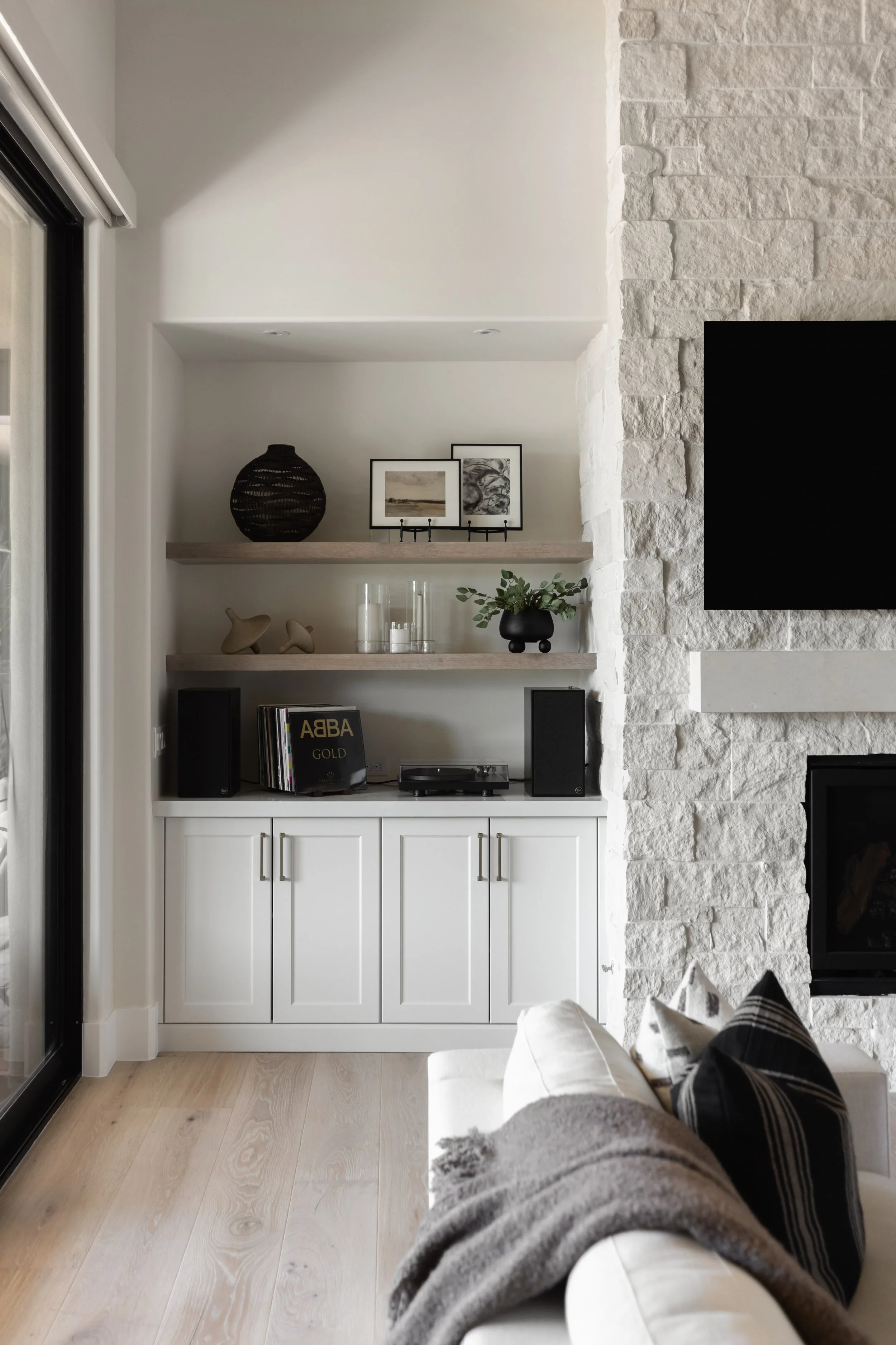 Living room with a white cabinet, decorated shelves with artwork, a black vase with greenery, speakers, a turntable, and a TV on a stone wall. Part of a person's legs and a blanket are visible in the foreground.