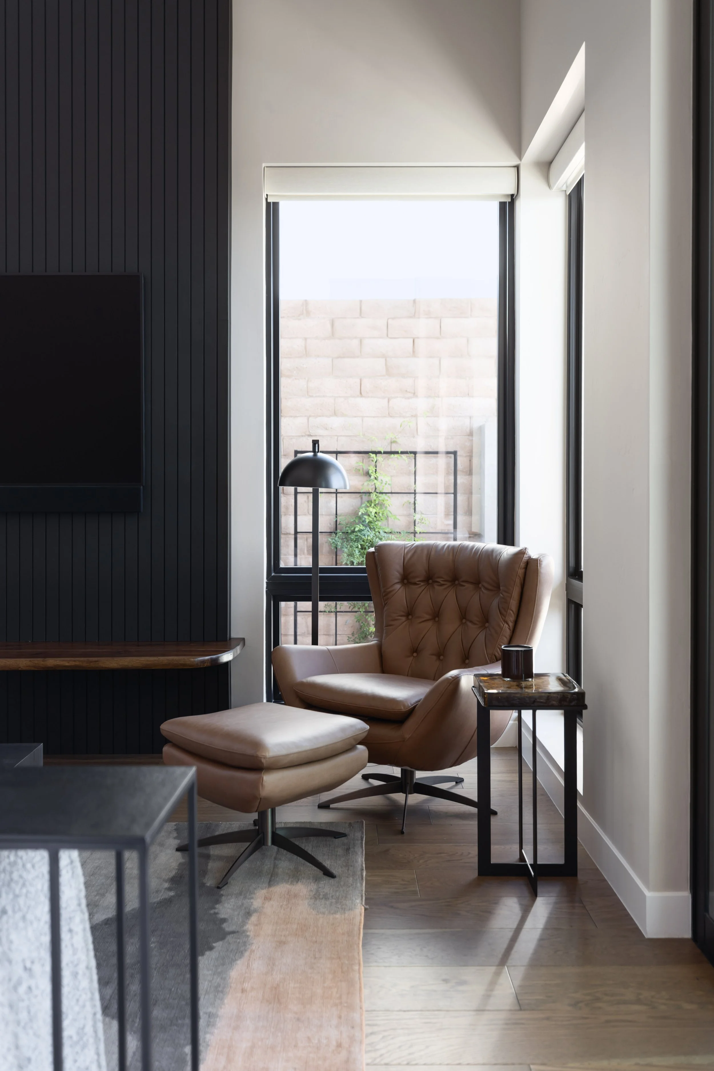 A cozy interior space with a large window, a tan armchair with a matching ottoman, a black floor lamp, and a side table with a candle holder. There's a black wall with vertical panels on the left and a beige rug on the floor.