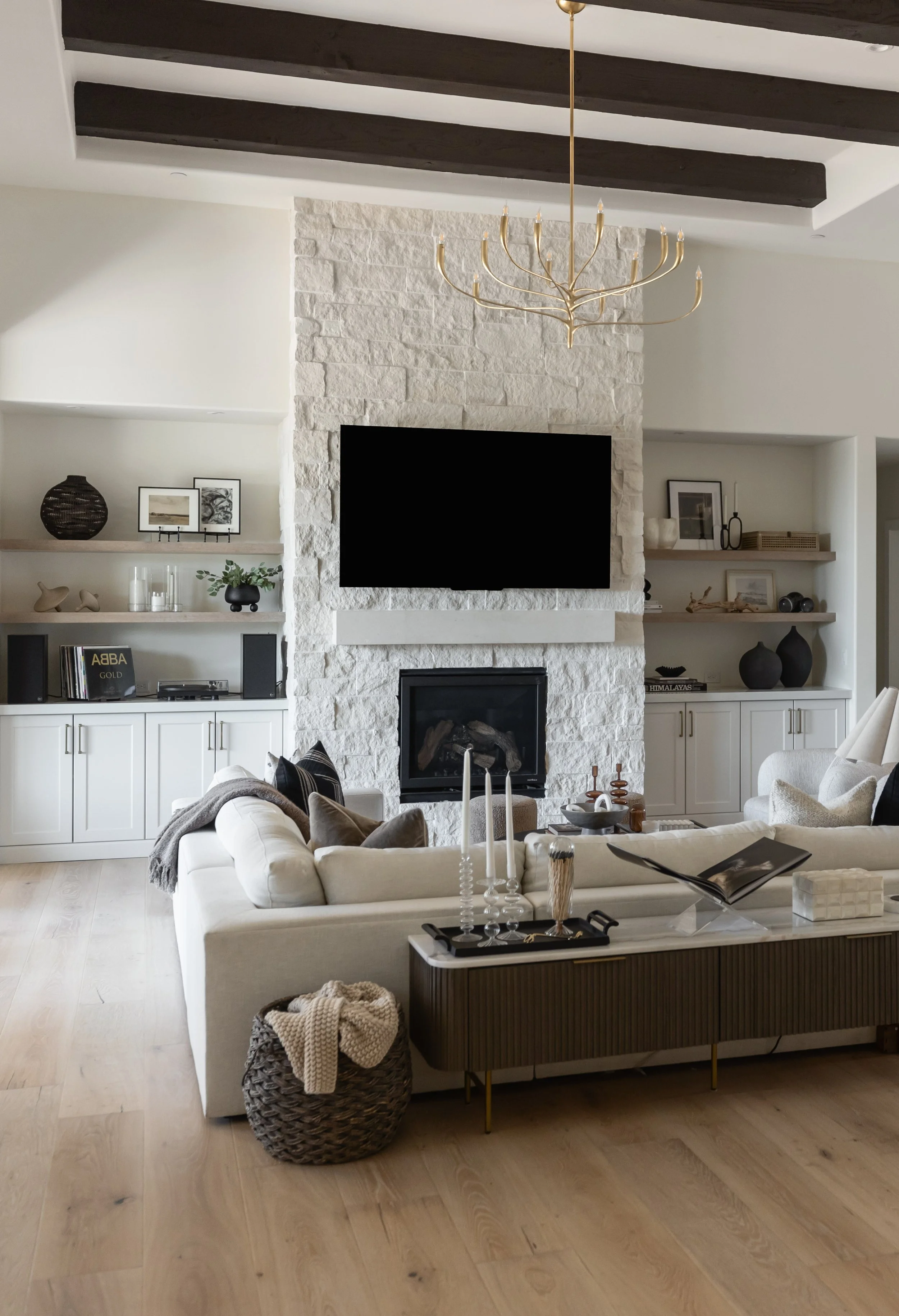 Modern living room with white brick fireplace, wall-mounted TV, beige sofa with pillows, wooden media console, two built-in shelves with decorative items, hardwood floor, and a gold chandelier.