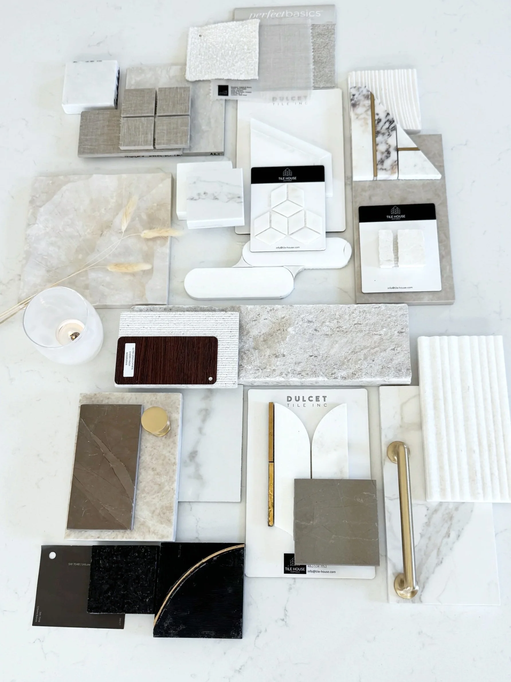 A variety of tile samples in different colors, patterns, and textures arranged on a white surface, along with a small lit candle, metal handles, and dried decorative grass.