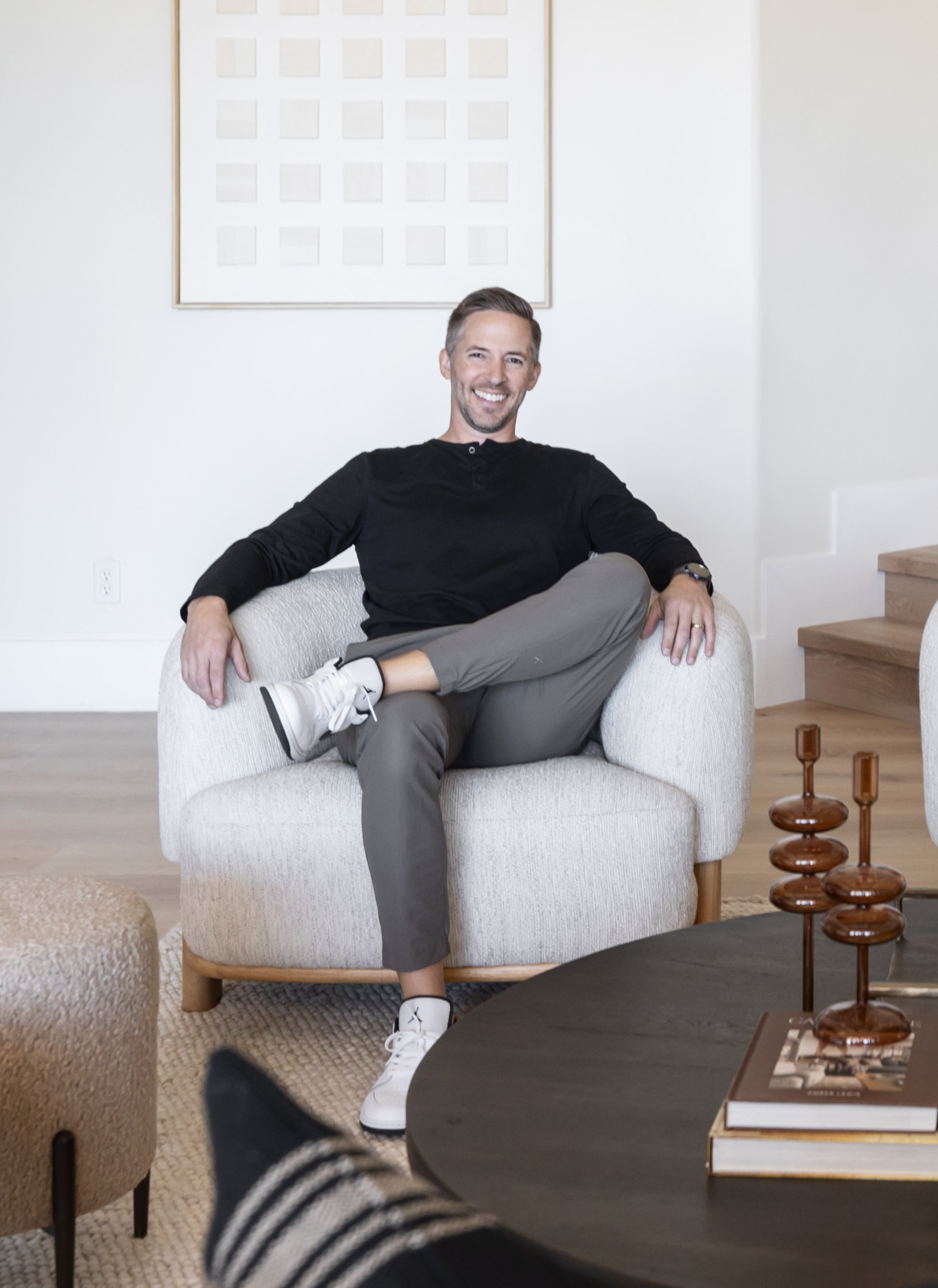 Man smiling and sitting on a light-colored armchair in a modern living room with a white wall, a wooden staircase, and decorative items on a coffee table.
