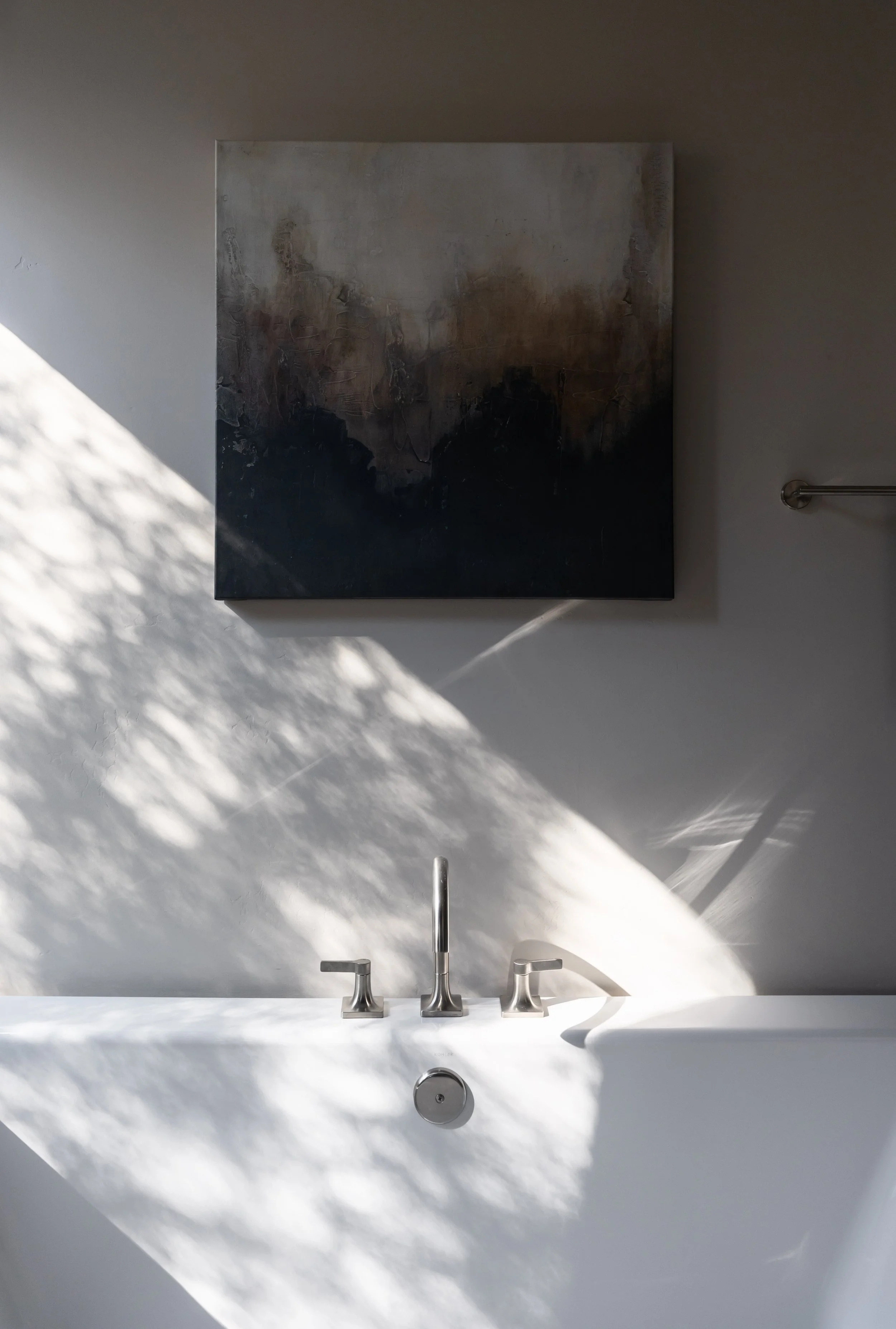 A white bathtub with a silver faucet and handles, against a gray wall with a large abstract painting and shadow patterns.