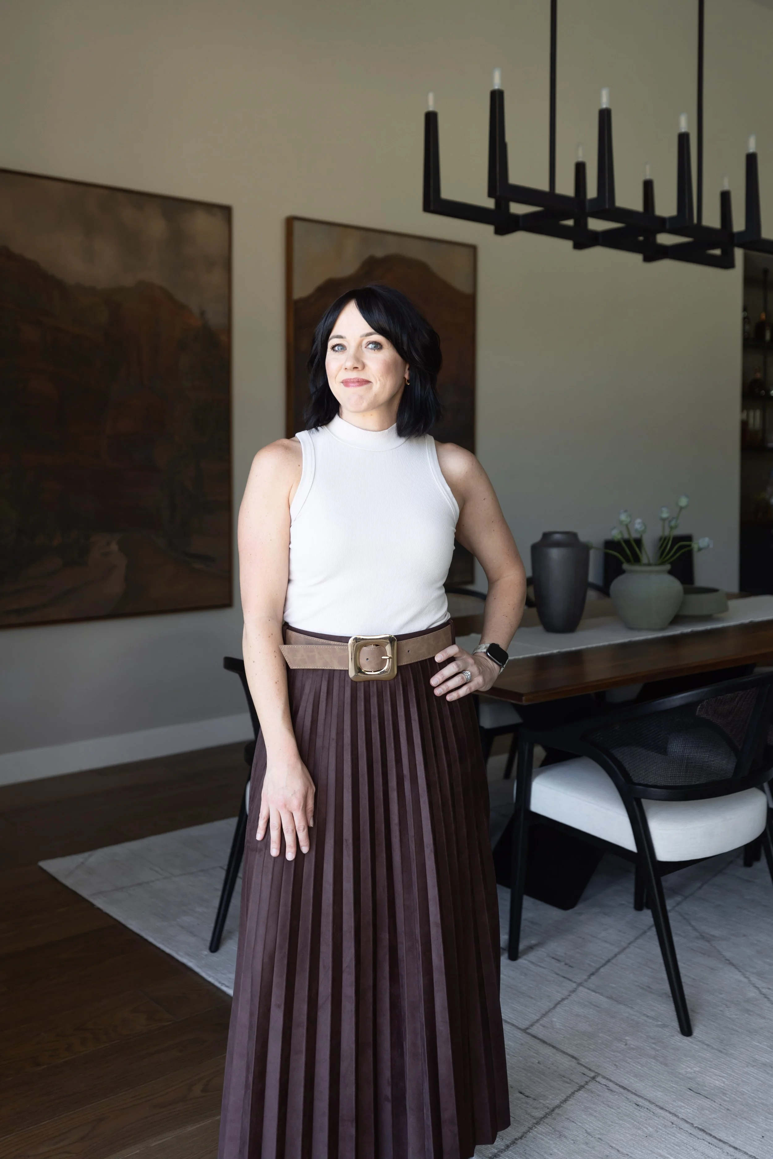 A woman with black hair and light skin standing in a dining room, wearing a sleeveless white top and a long pleated brown skirt, with paintings on the wall and a modern chandelier overhead.