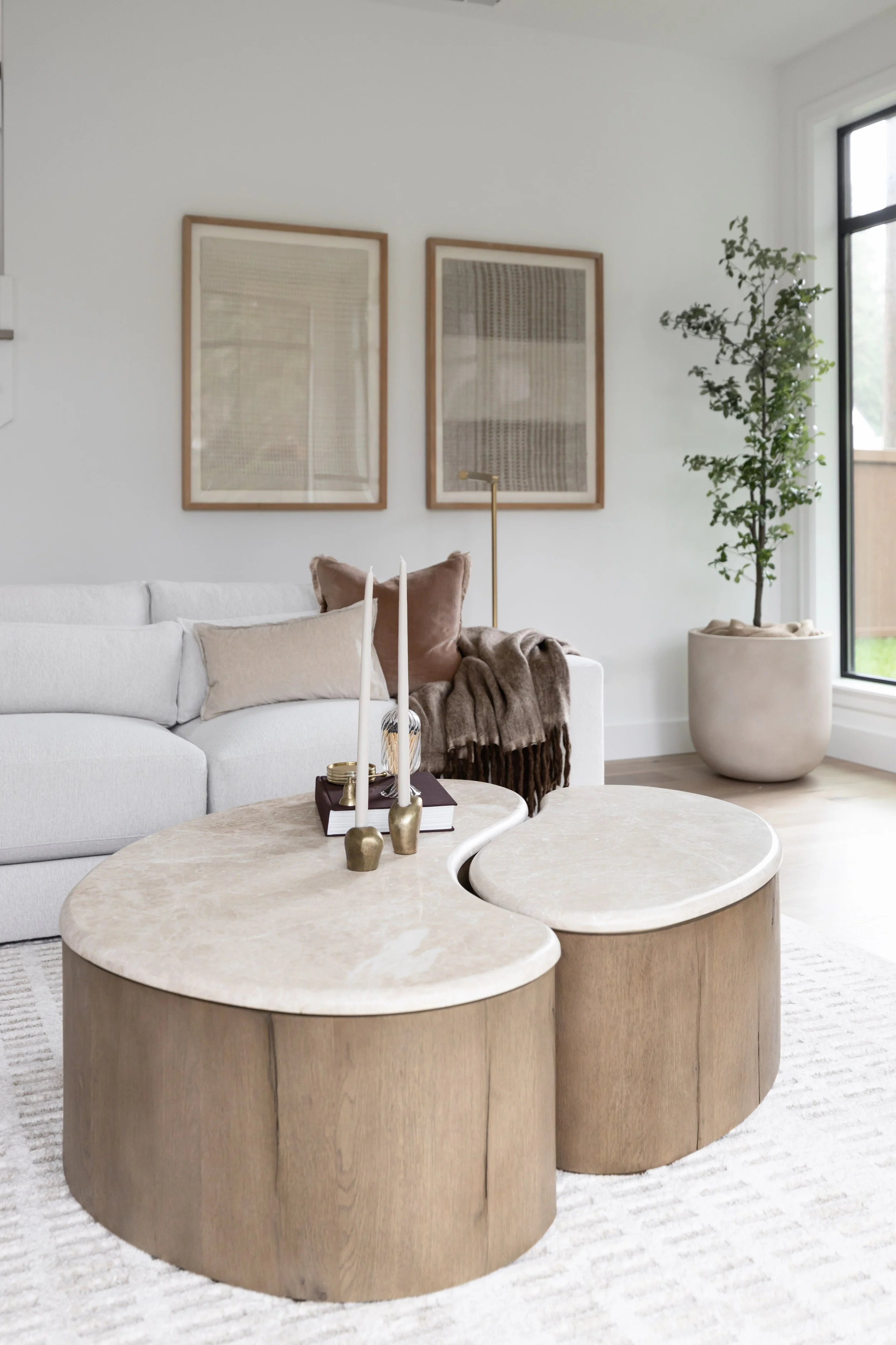 Living room with two wooden oval coffee tables, beige sofa with pillows, wall art, and a potted plant near a large window.