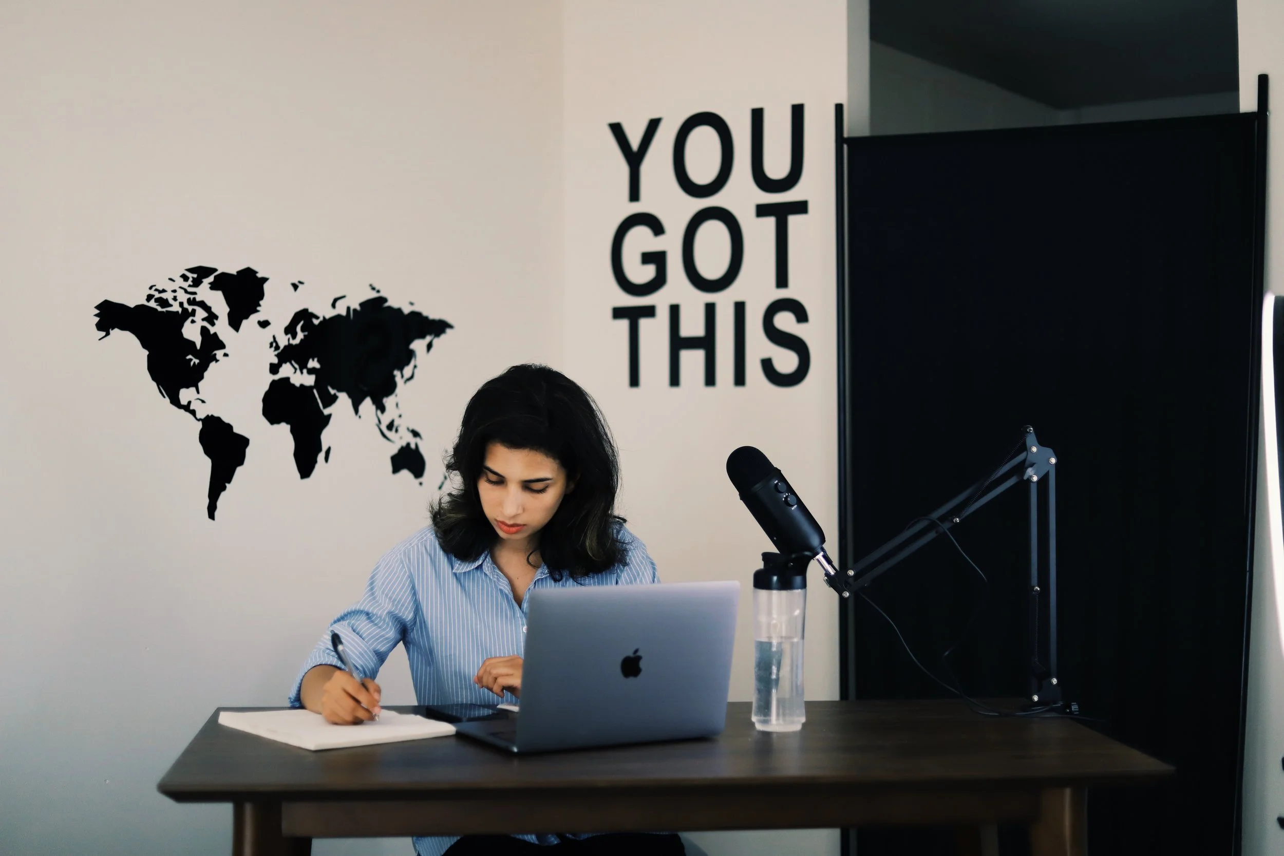 A woman sitting at a desk, writing in a notebook, with a laptop, a water bottle, and a microphone. Behind her is a wall with a world map decal and the phrase 'YOU GOT THIS'.