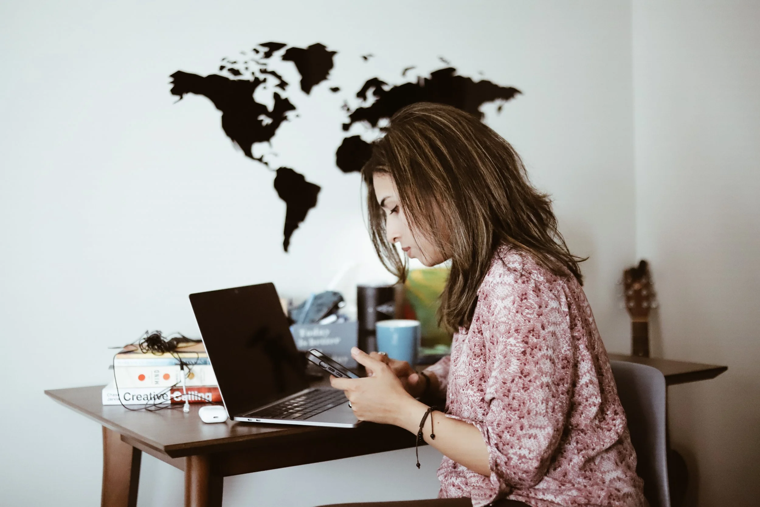 Woman with brown hair using her smartphone at a wooden desk with a laptop, books, a coffee mug, and a guitar in the background, with a black world map wall decal.