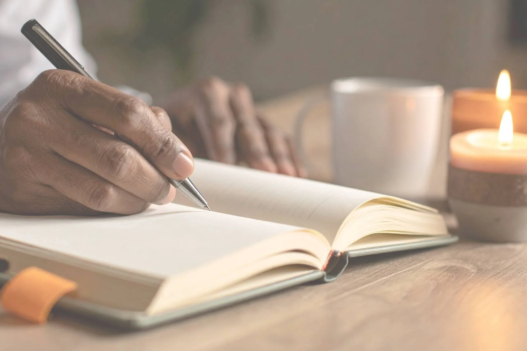 Persons hand holding pen journaling in open blank book, with candle and a mug depicting January reflection encouraging slow living and emotional wellbeing