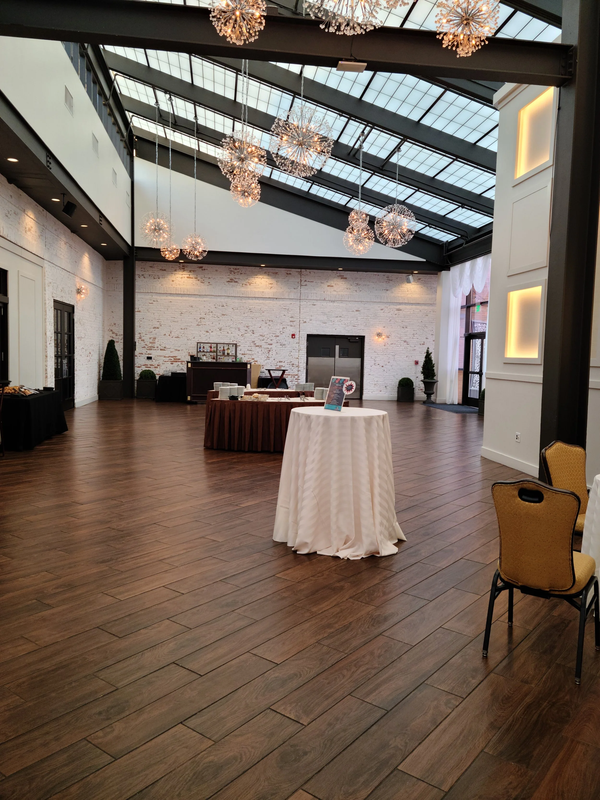 Empty banquet hall with wooden floors, white brick walls, round tables with tablecloths, and hanging chandeliers in an industrial-style space with a glass ceiling.