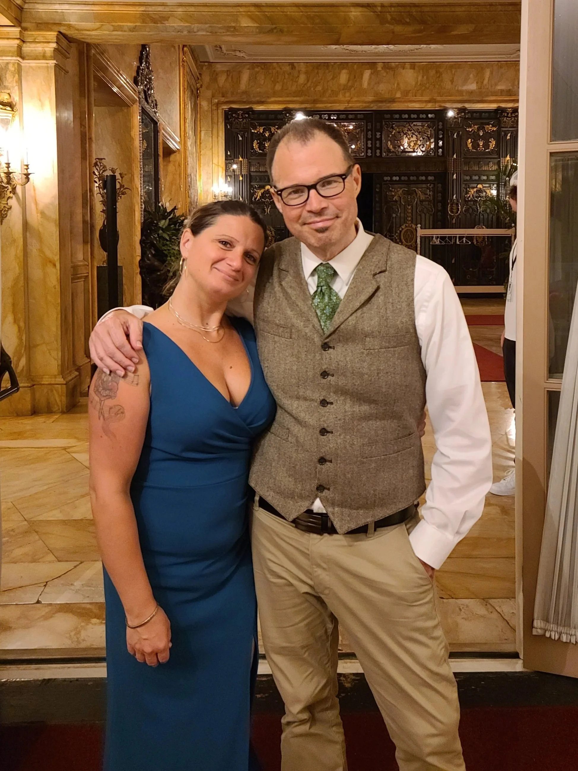 A man and woman standing together, smiling, in an elegant indoor setting. The woman has dark hair, tattoos on her arm, and is wearing a blue dress. The man has glasses, a beard, and is dressed in a white shirt, gray vest, green tie, and beige pants.