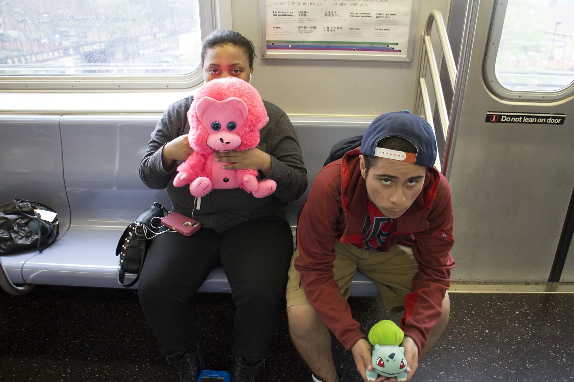 A young woman sitting on public transit holding a pink plush gorilla toy, and a young man kneeling on the floor holding a Bulbasaur plush toy with a green bulb on its back.