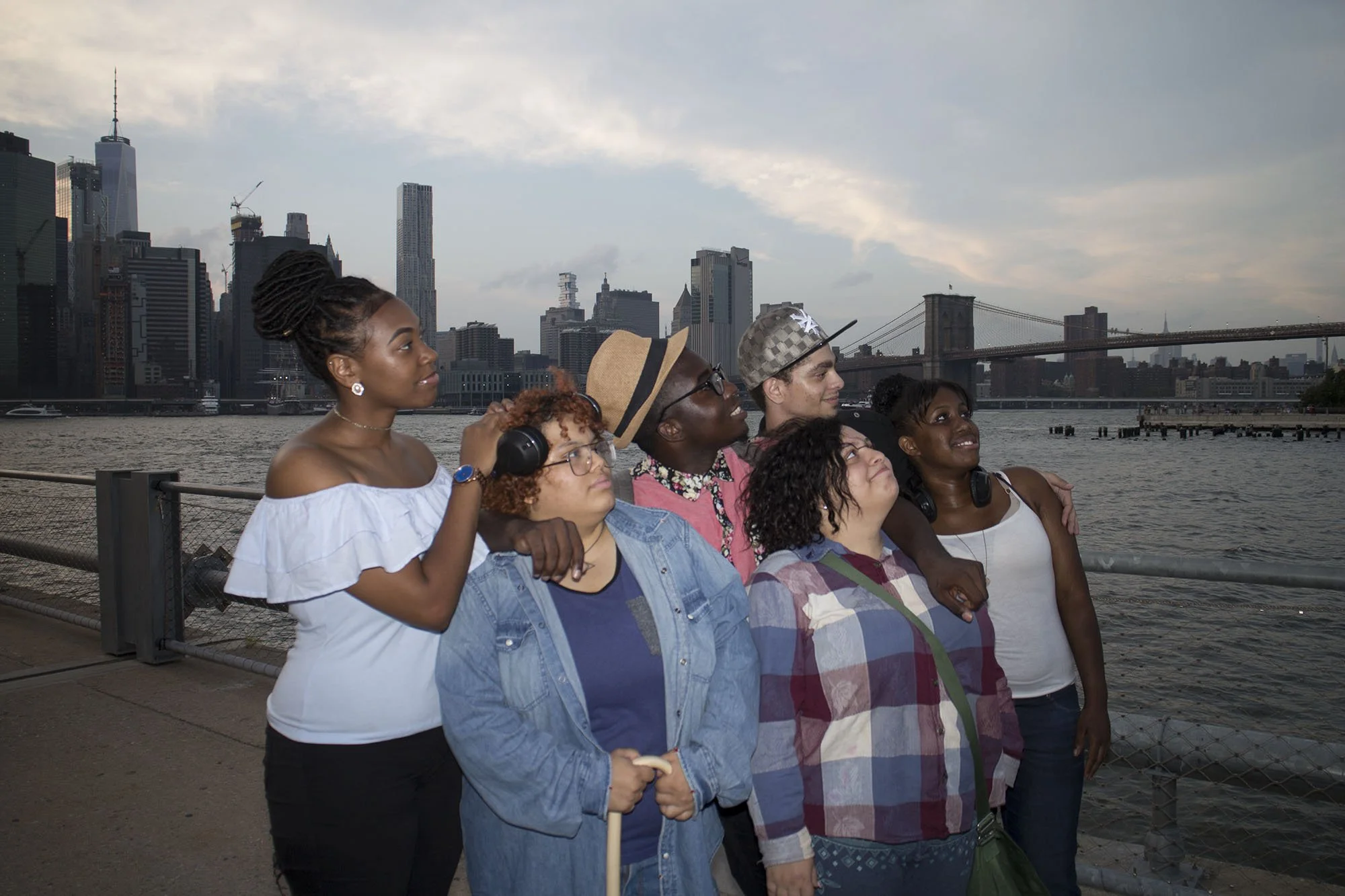 Group of six diverse friends sightseeing by the water in New York City, with the Brooklyn Bridge and Manhattan skyline in the background.