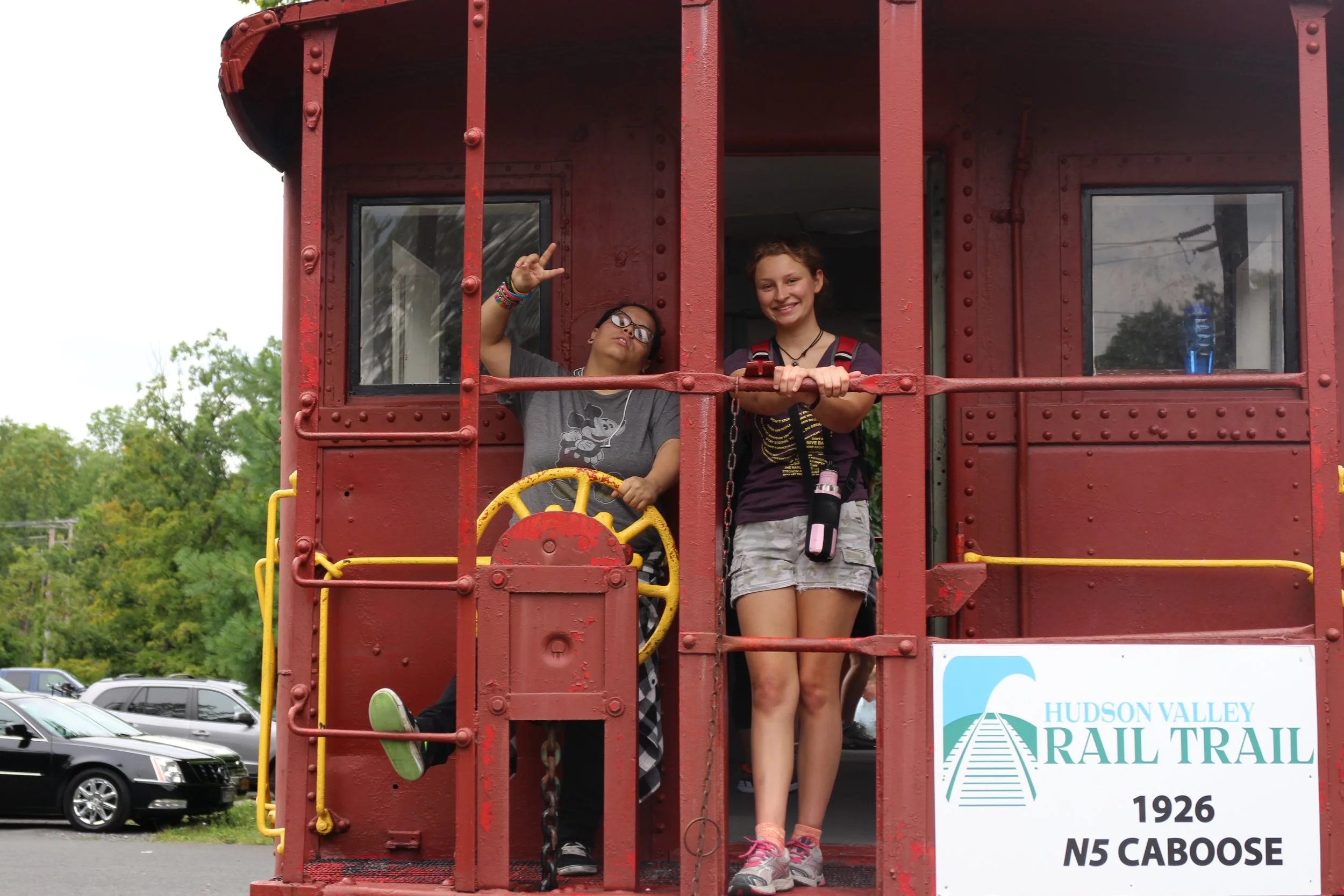 Two young women on a red historic railroad caboose car, one making a peace sign and the other smiling, at the Hudson Valley Rail Trail.