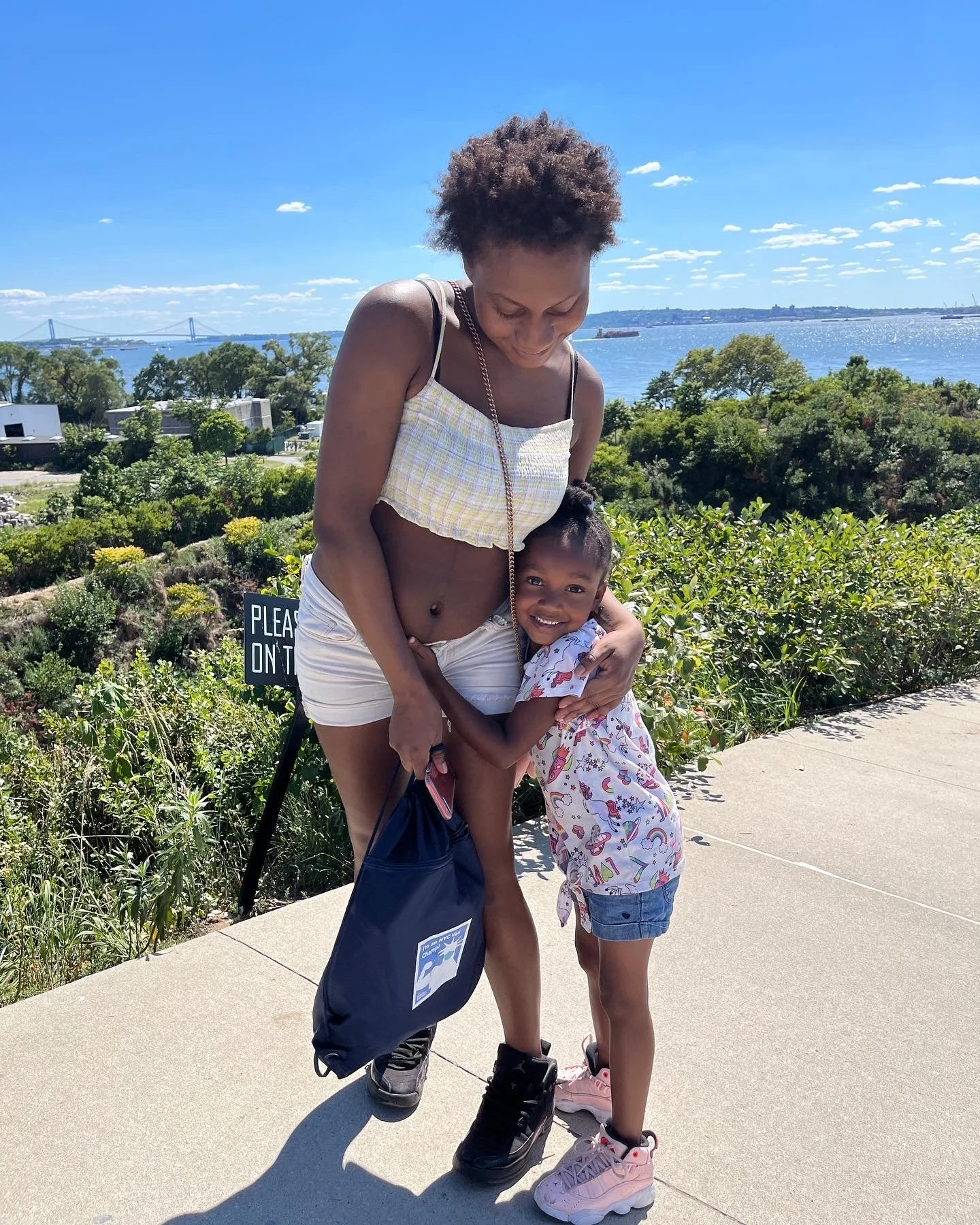 A woman and a young girl are hugging on a sidewalk beside greenery with a river and blue sky in the background. The woman is wearing a light yellow checkered crop top and white shorts, and the girl is wearing a colorful t-shirt, denim shorts, and pin