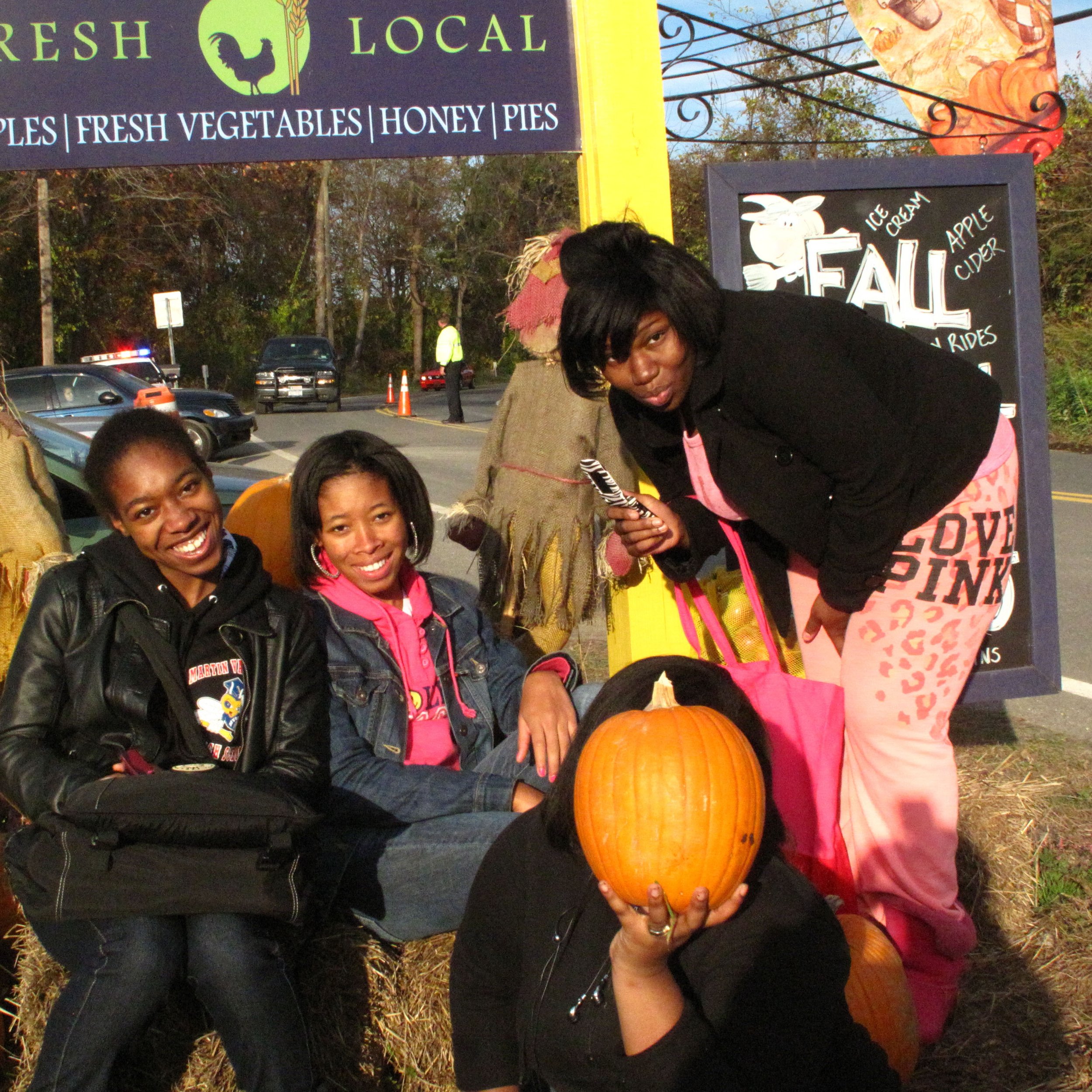Group of four women wearing casual clothes, two of them sitting on hay bales and one standing, posing with Halloween pumpkins in front of a fall-themed outdoor display.
