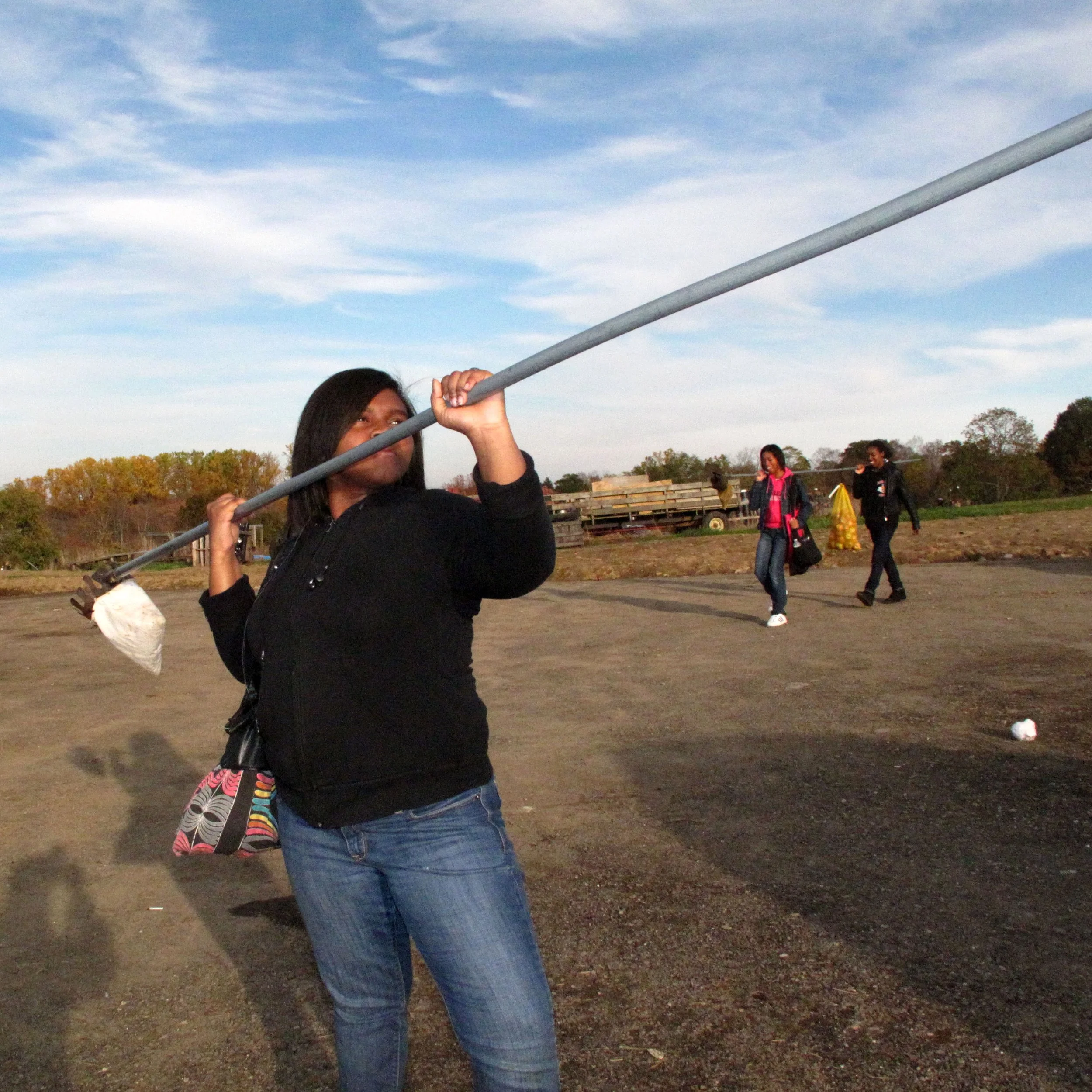 A woman holding a long pole with a bag at the end, standing outdoors on a dirt field during daytime, with three other people walking in the background.