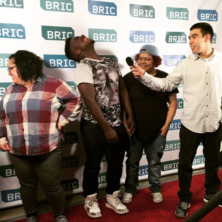 Group of four people standing in front of a BRIC backdrop. They are smiling and laughing; one person is holding a microphone near another's face, suggesting an interview or a fun event.
