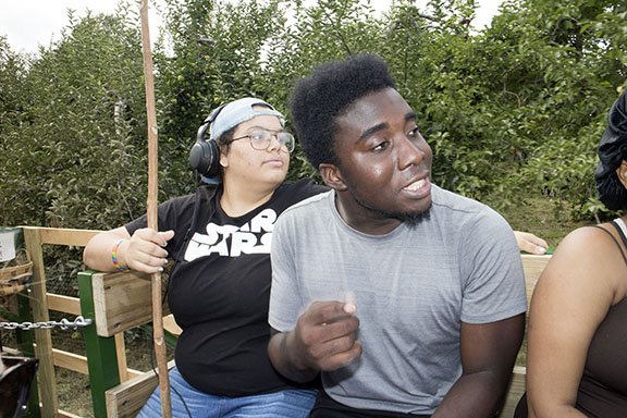 Two people sitting outdoors next to a green hedge, one wearing headphones and a Star Wars shirt, the other wearing a gray t-shirt and gesturing with their hand.