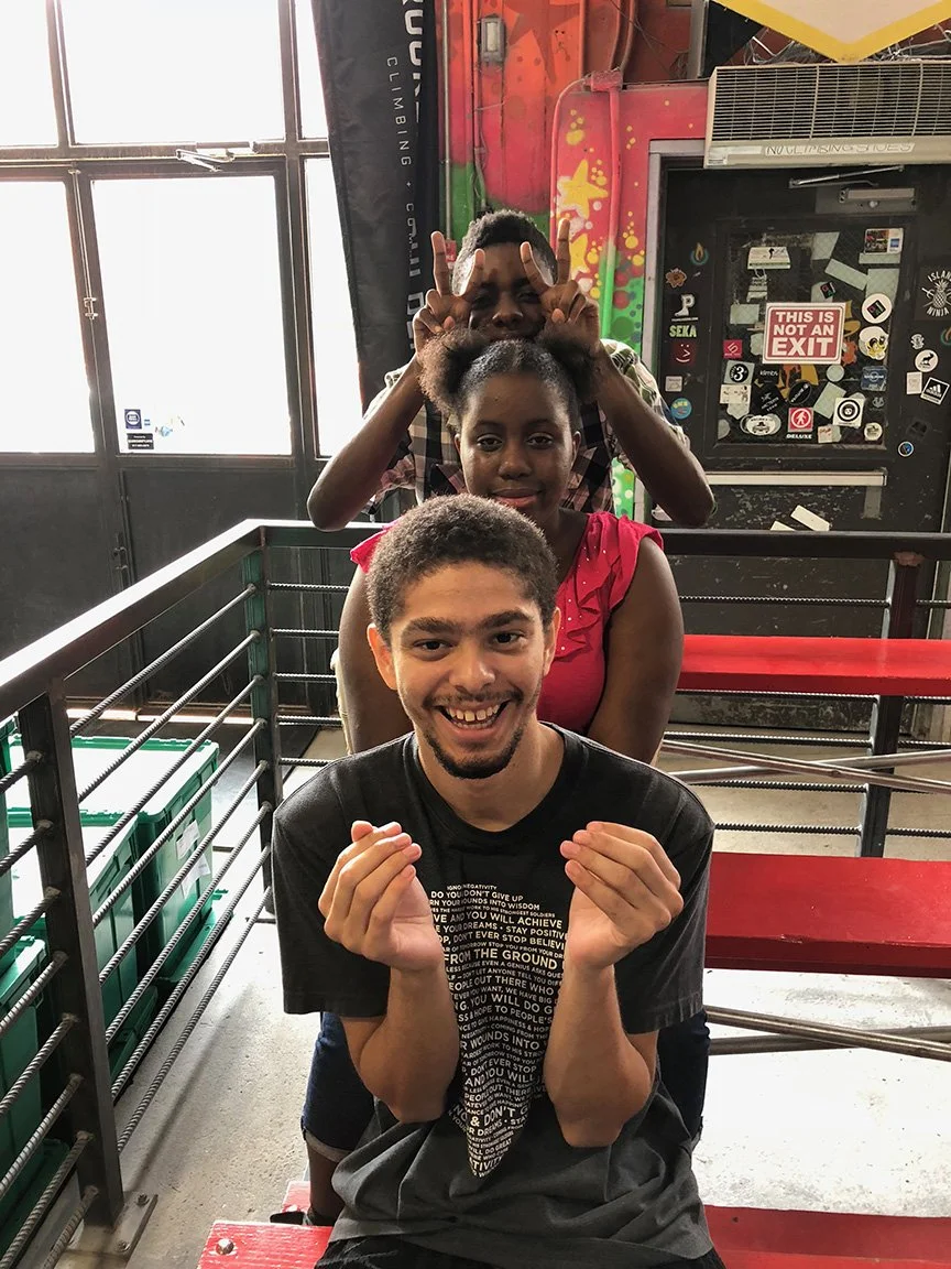 Three smiling young people in a colorful indoor setting, two males and one female, with one male in front making a finger gesture and a male behind making a peace sign with his fingers above the woman's head.
