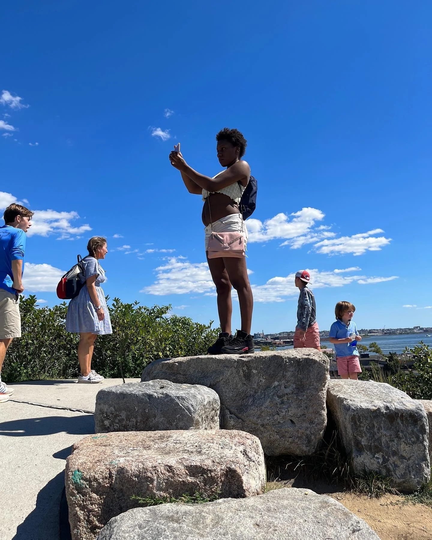 A group of people outdoors on a rocky area under a bright blue sky with scattered clouds. One woman is standing on a large rock, taking a photo or video with her phone. Several children and a man are nearby, some standing on the ground and others on 