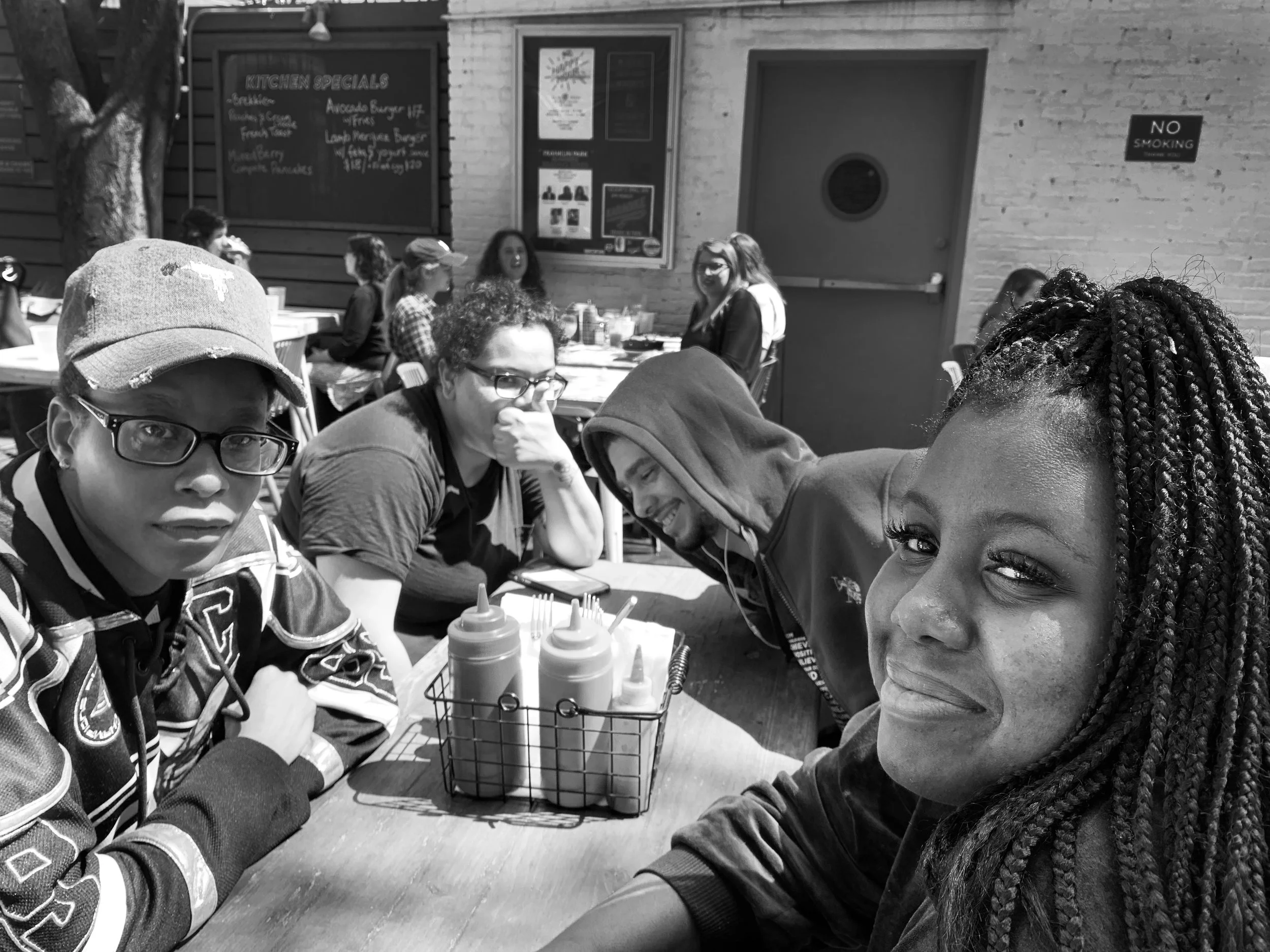 Group of five young adults sitting at an outdoor restaurant table, with condiment bottles in a wire basket in the foreground. The woman in the foreground has braided hair and is smiling slightly. The others are engaged in conversation or looking at t