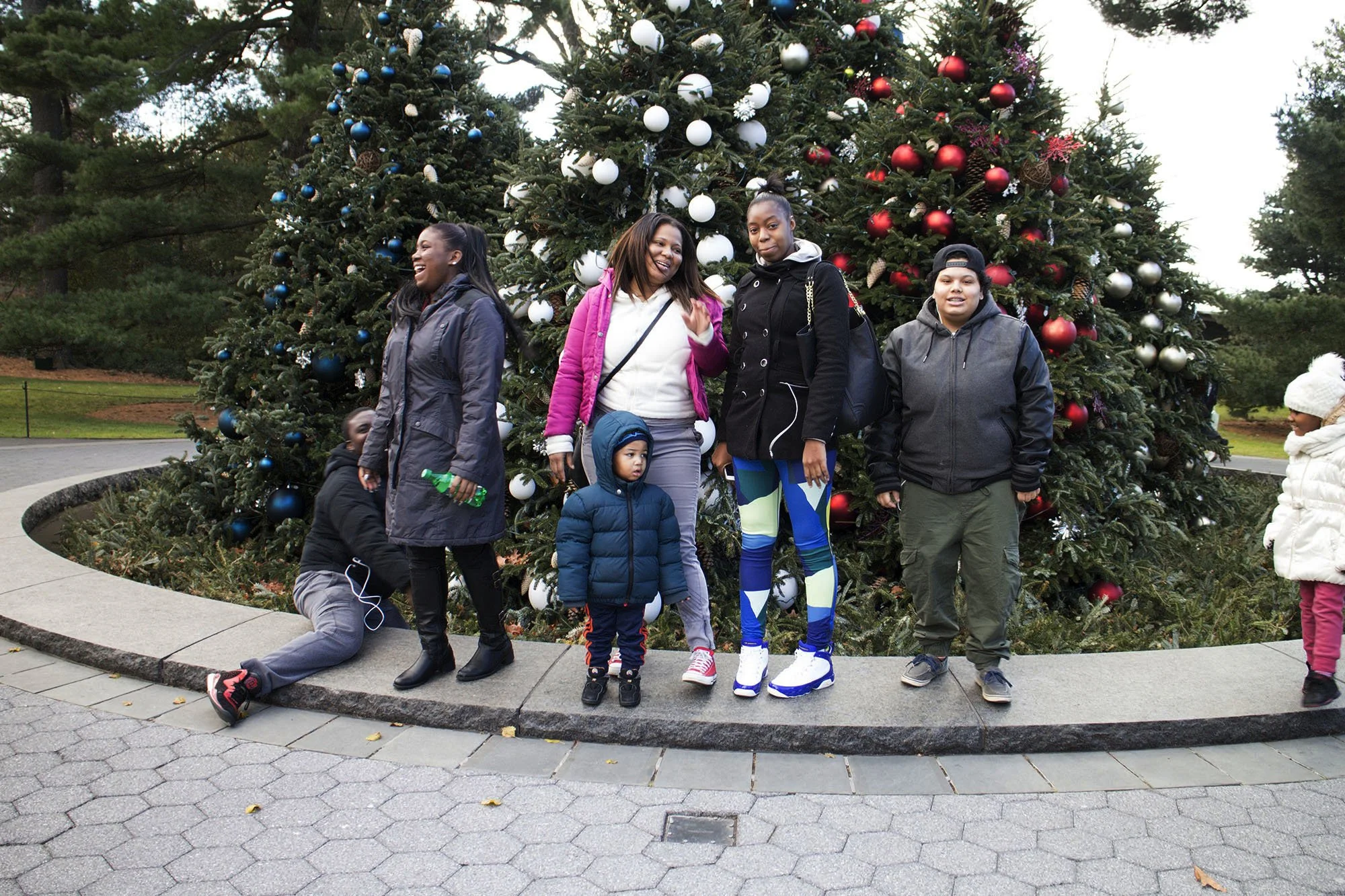 Group of people, including children and adults, standing in front of decorated Christmas trees outdoors during the daytime.