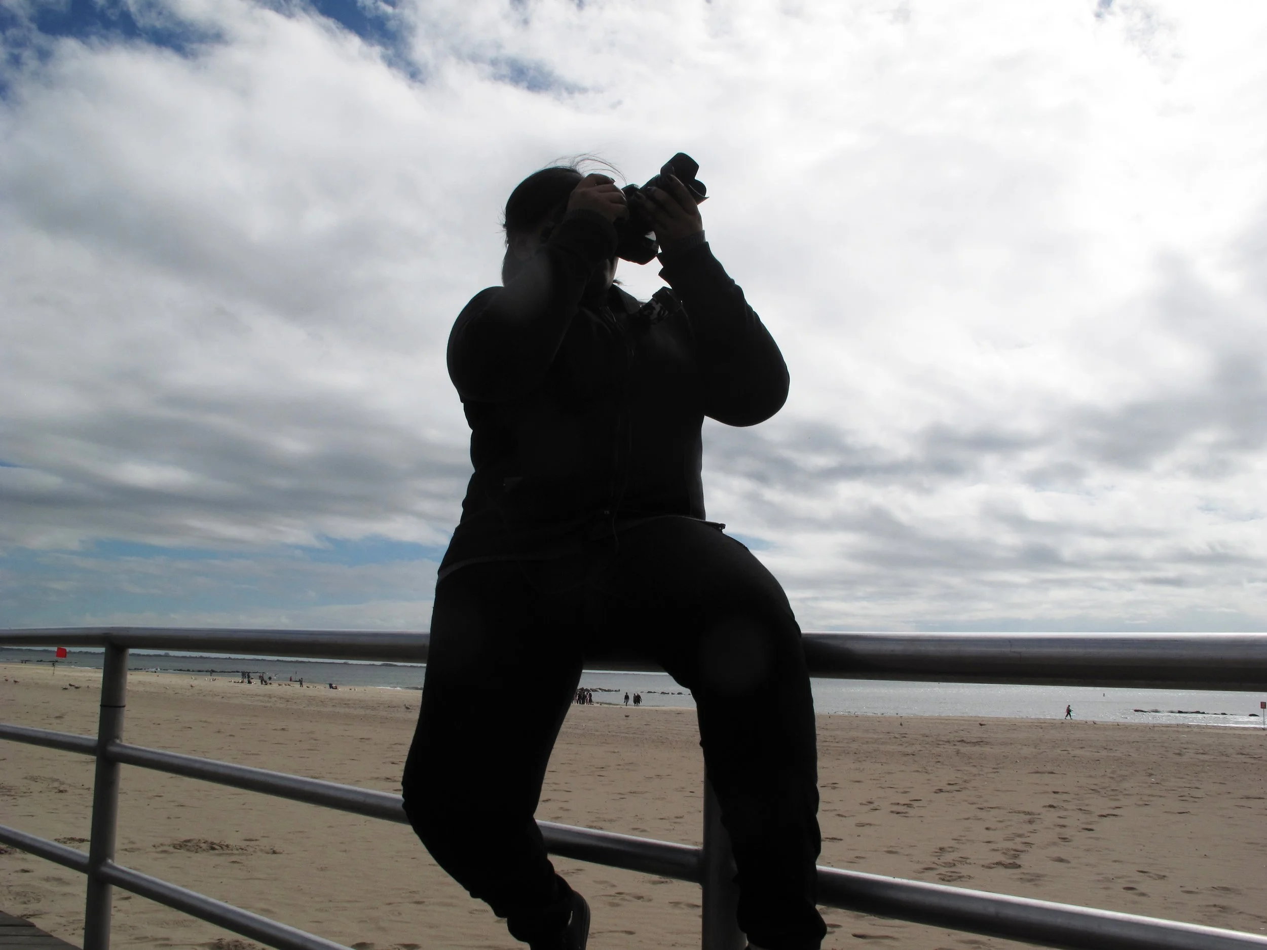 Silhouette of a person taking a photograph with a camera on a beach, with cloudy sky in the background.