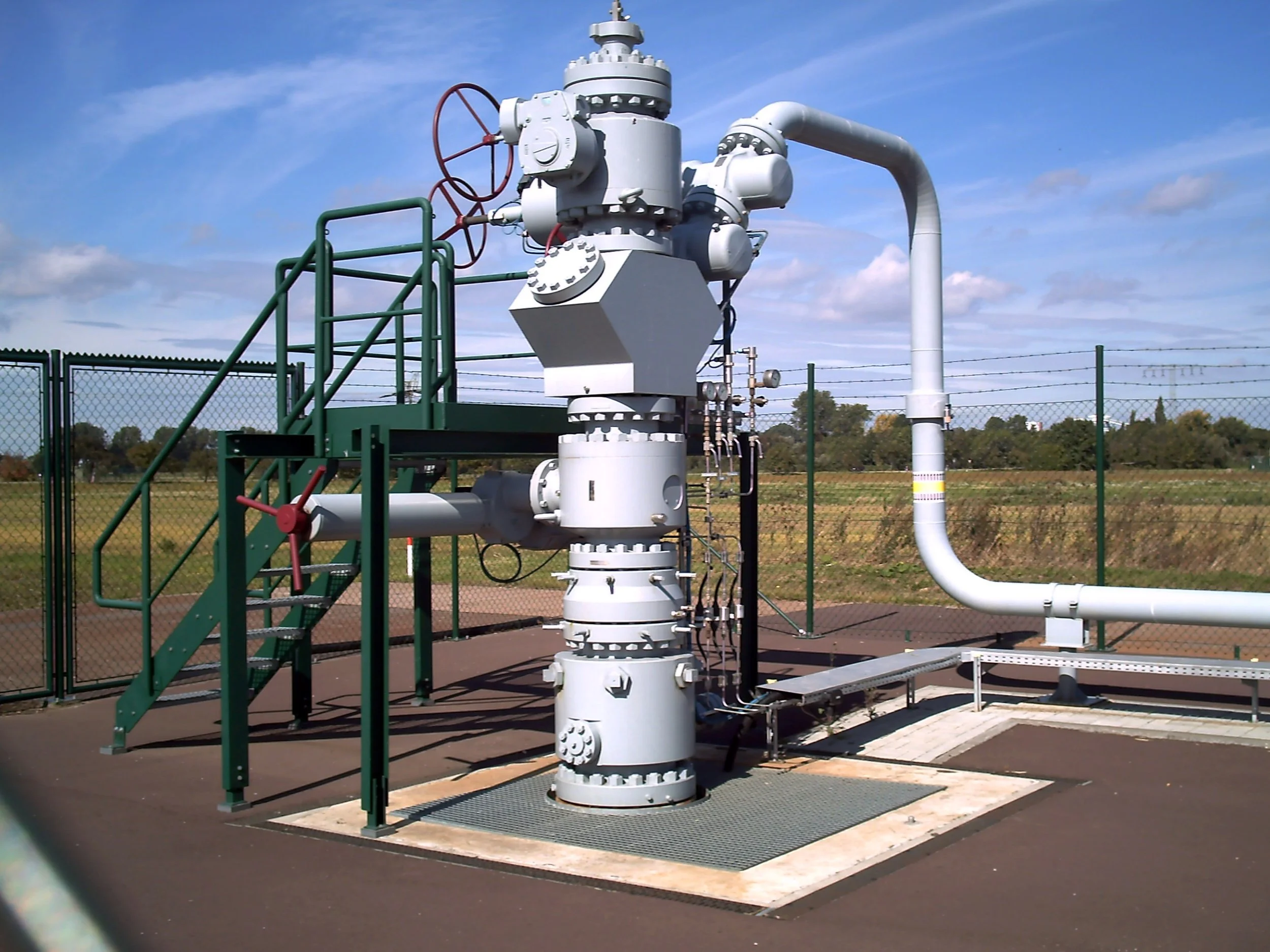 An outdoor industrial valve system with pipes, gauges, and red and green handwheels, surrounded by a chain-link fence and set against a landscape with a partly cloudy sky.