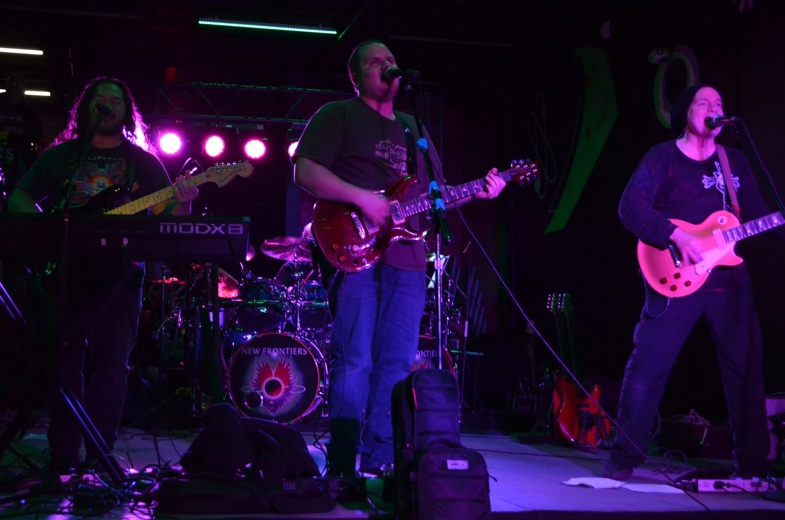 A band performing on stage with three musicians playing guitars, purple and green stage lighting, and a drum set with a "New Frontiers" logo in the background.