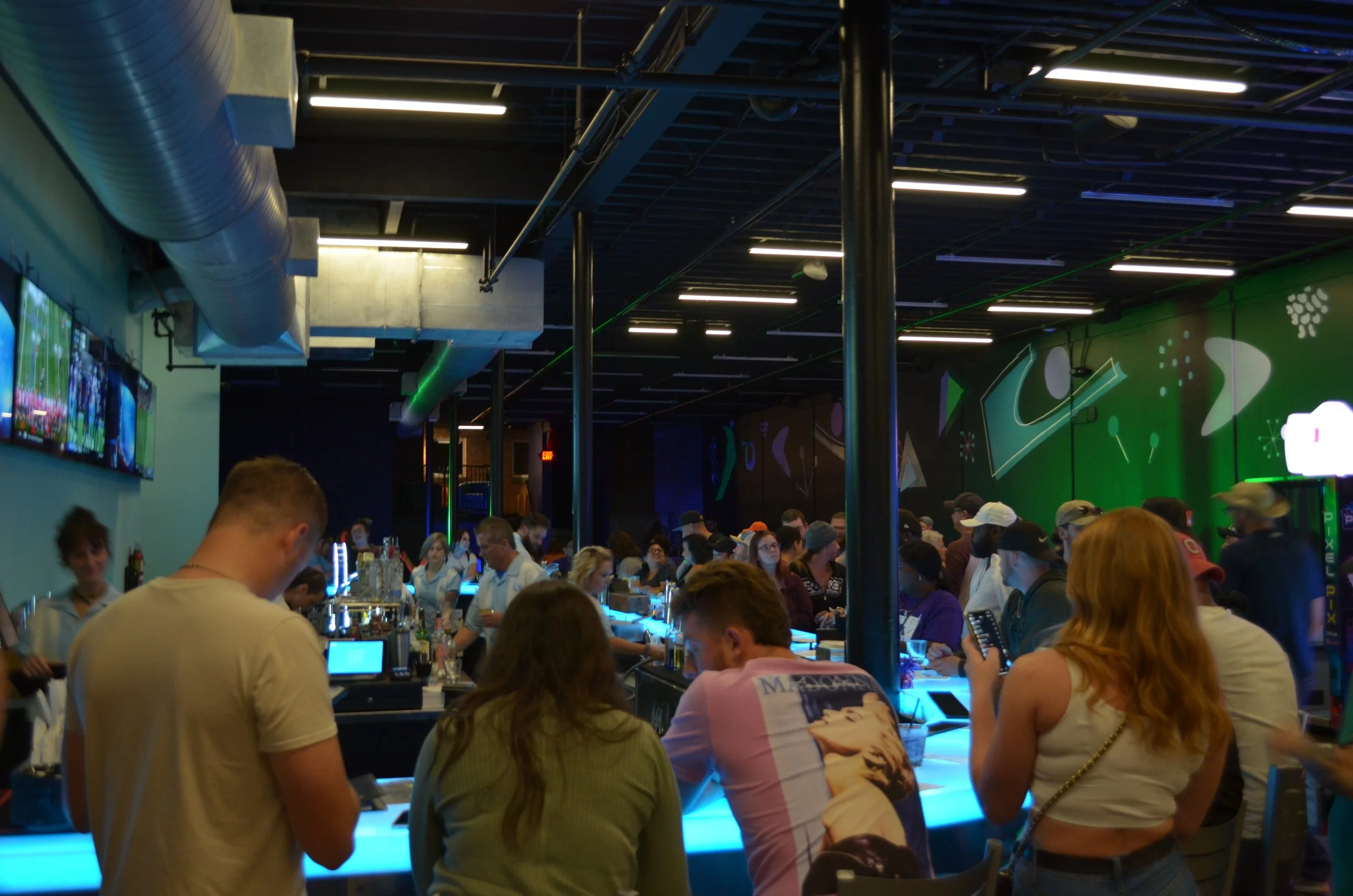 Crowd of people at a neon-lit bar or nightclub with a green abstract mural on the wall.